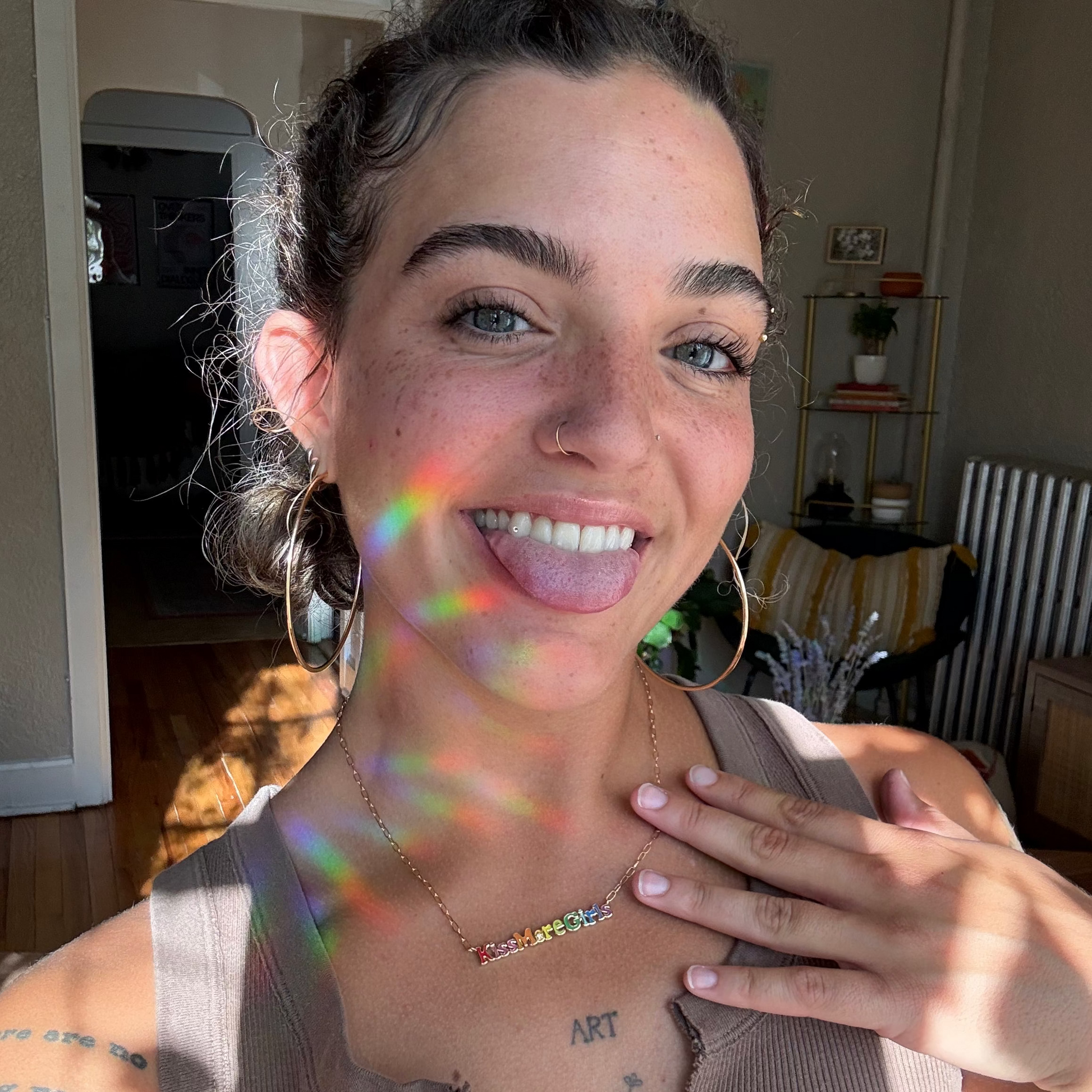 A young woman with blue eyes and hoop earrings smiles, sticking out her tongue as rainbow light reflects on her face. She wears a tank top, the Kiss More Girls Necklace from Local Eclectic, and stands in a cozy, sunlit room.