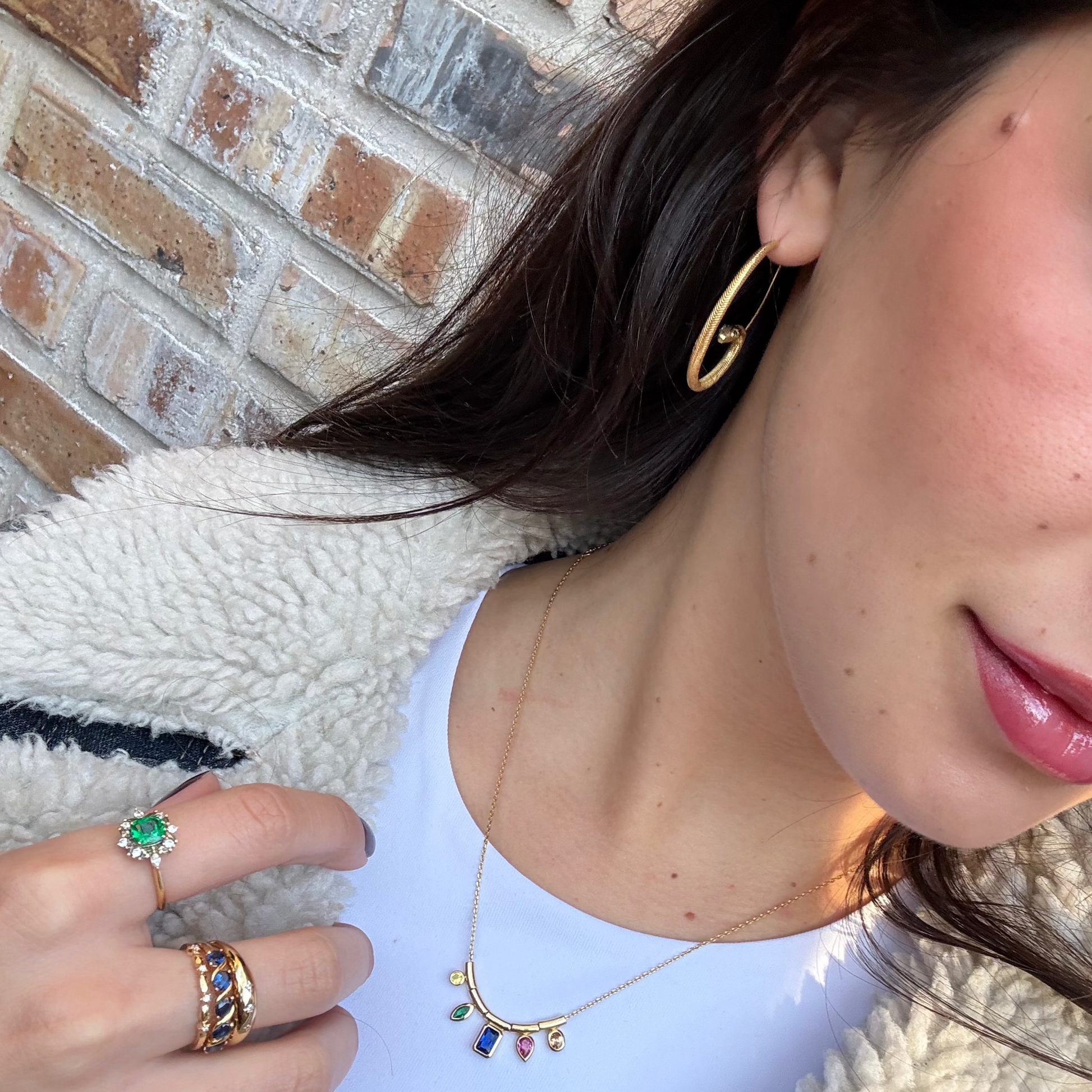 A woman in a white top and fuzzy jacket poses against a brick wall, wearing Mini Snake Hoops gold earrings, a gemstone necklace, and several rings including one with a large green stone.