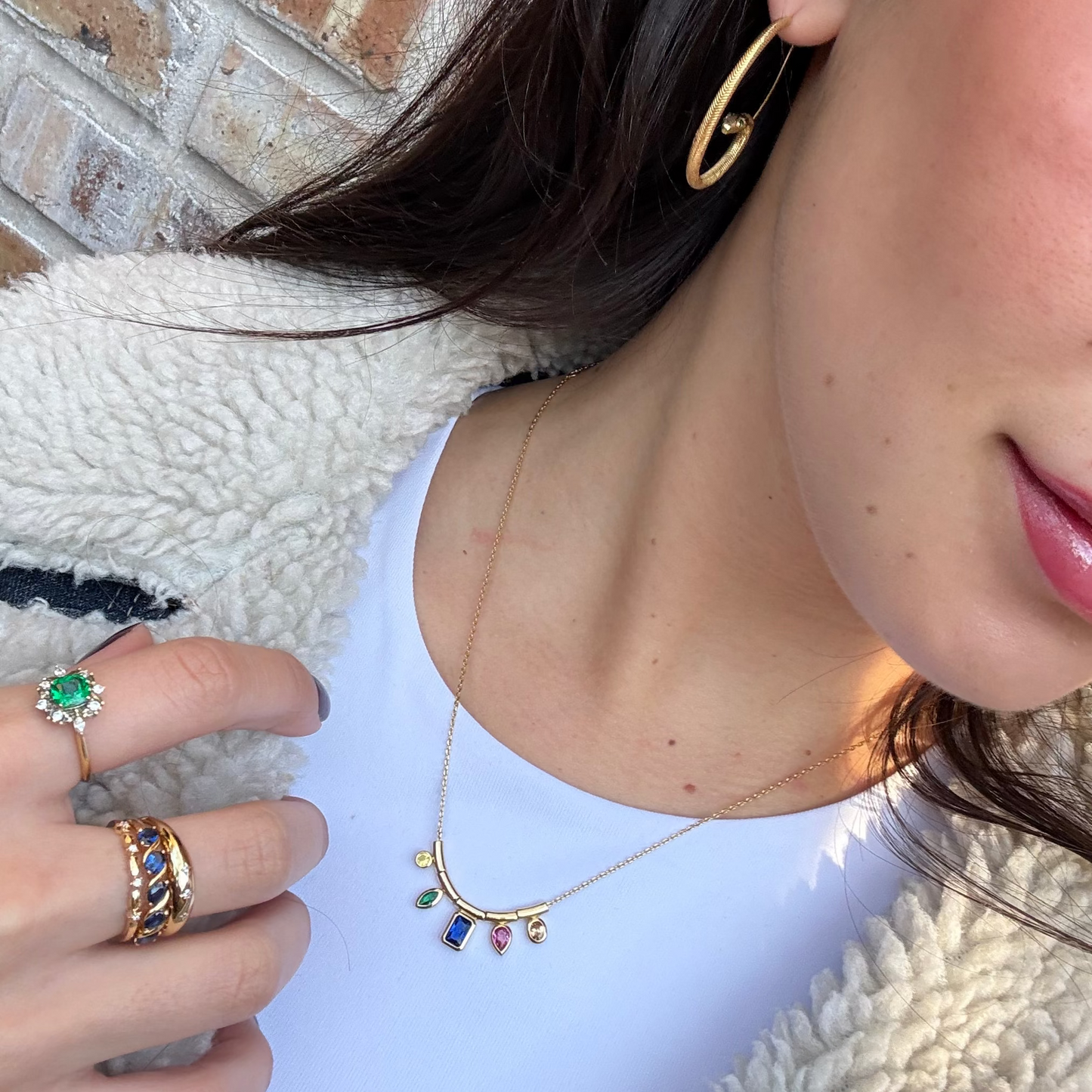 A woman wears the Technicolor Gem Drop Necklace, gold hoop earrings, and rings with green, blue, and purple stones. She pairs her jewelry with a white top and a textured cream jacket.
