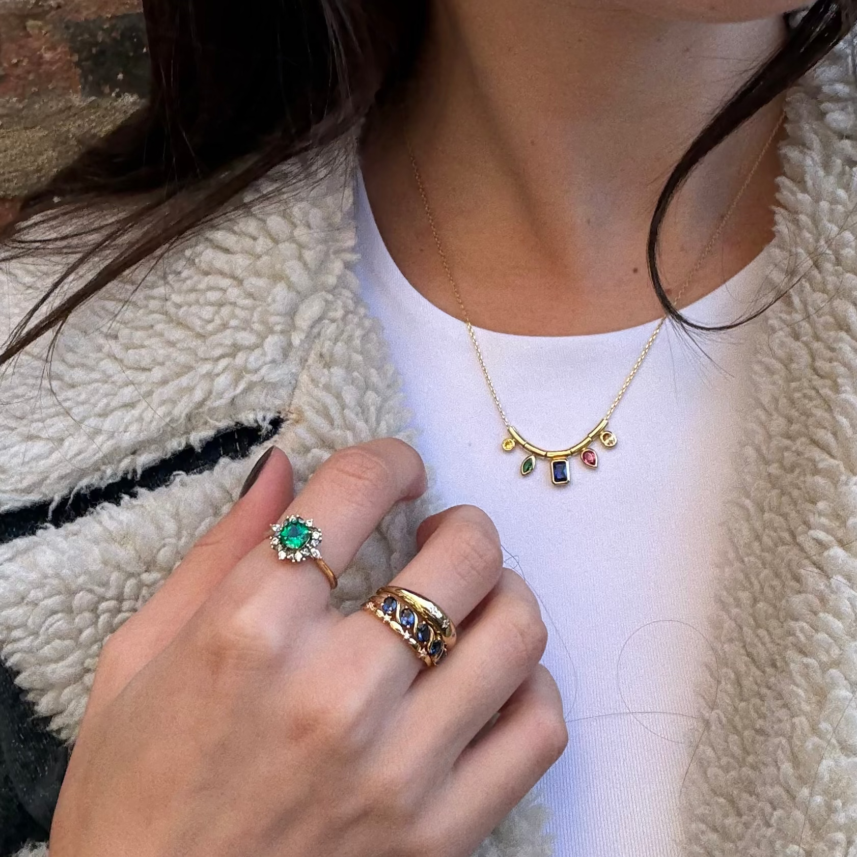 A person in a white top and fuzzy jacket showcases a gold necklace with colorful charms, a green gemstone ring, and the Starry Stacking Band—gold rings set with blue and white stones.