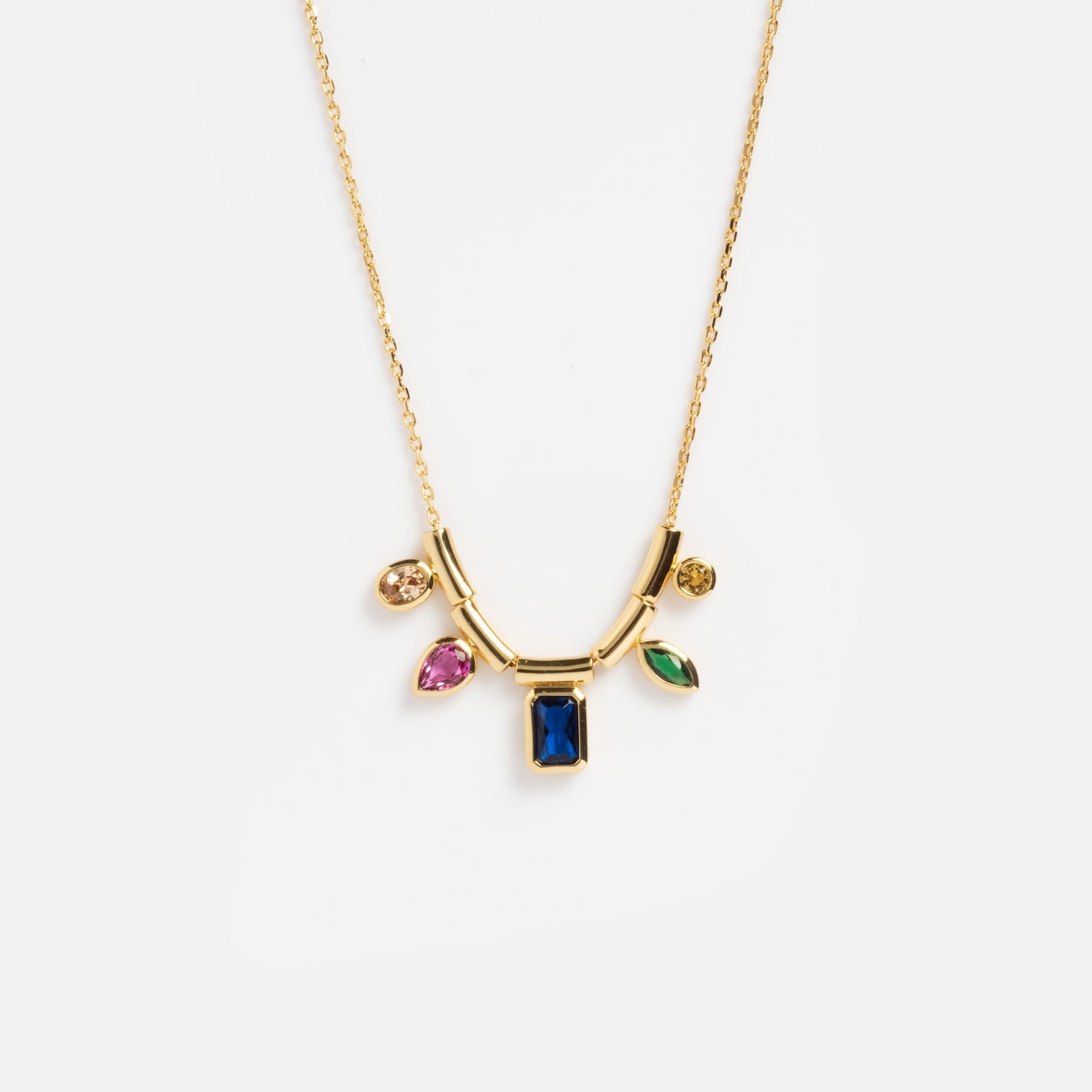 The Technicolor Gem Drop Necklace features a fine gold chain with a central rectangular blue gemstone pendant and four dangling charms in pink, green, and yellow gemstones.
