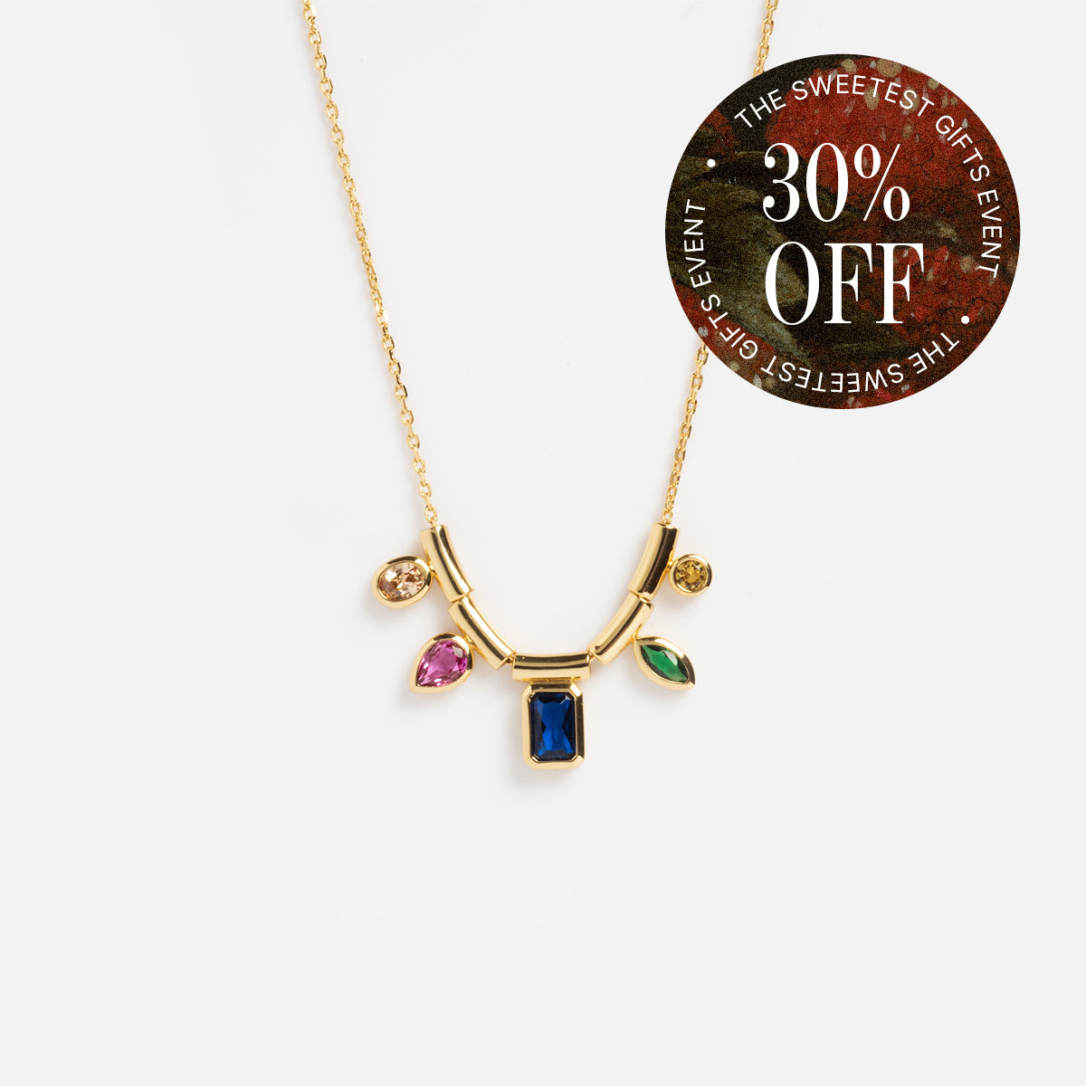 The Technicolor Gem Drop Necklace, yellow gold plated and embellished with multicolored gem charms, is shown on a white background. A round sticker at the top right says “30% OFF - THE SWEETEST GIFTS EVENT” in white on a patterned backdrop.