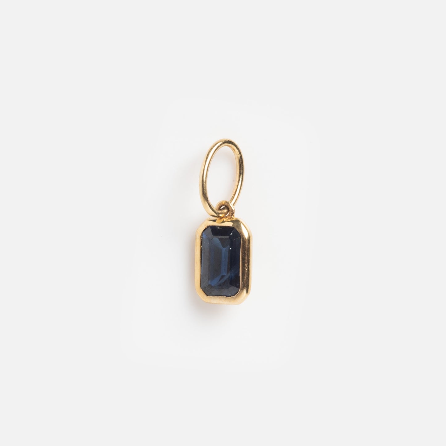 A Solid Gold Sapphire Baguette Charm Sample featuring a small gold pendant with a rectangular dark blue sapphire, attached to a simple gold loop and shown on a plain white background.
