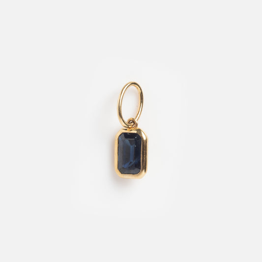 A Solid Gold Sapphire Baguette Charm Sample featuring a small gold pendant with a rectangular dark blue sapphire, attached to a simple gold loop and shown on a plain white background.