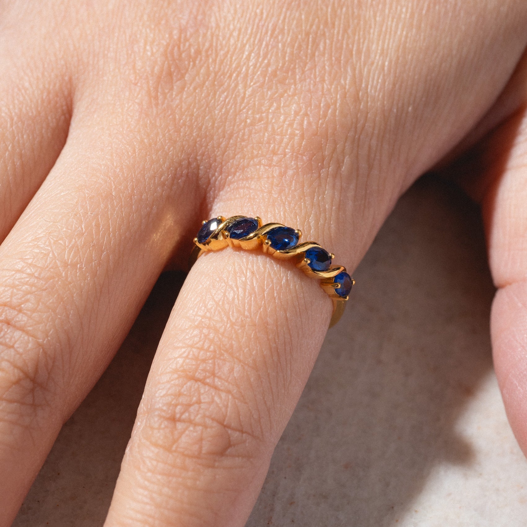 A hand wears the Blue Twist Vintage Inspired Ring, featuring a gold band with a row of small oval blue stones, shown in close-up against a neutral background.