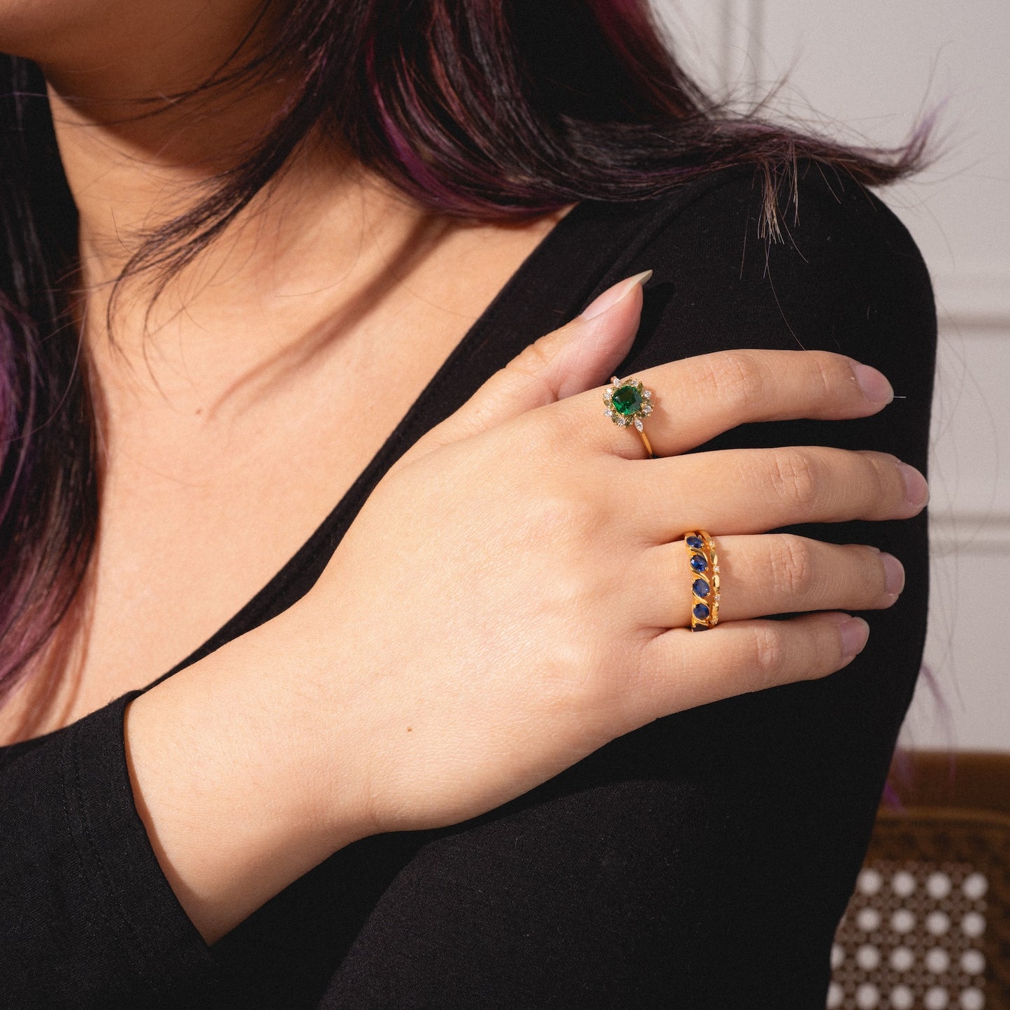 A woman with long hair and a black top rests her hand on her arm, showcasing the Green Gardens Vintage Inspired Ring featuring a large green stone surrounded by small gems, along with a gold band set with blue oval stones.