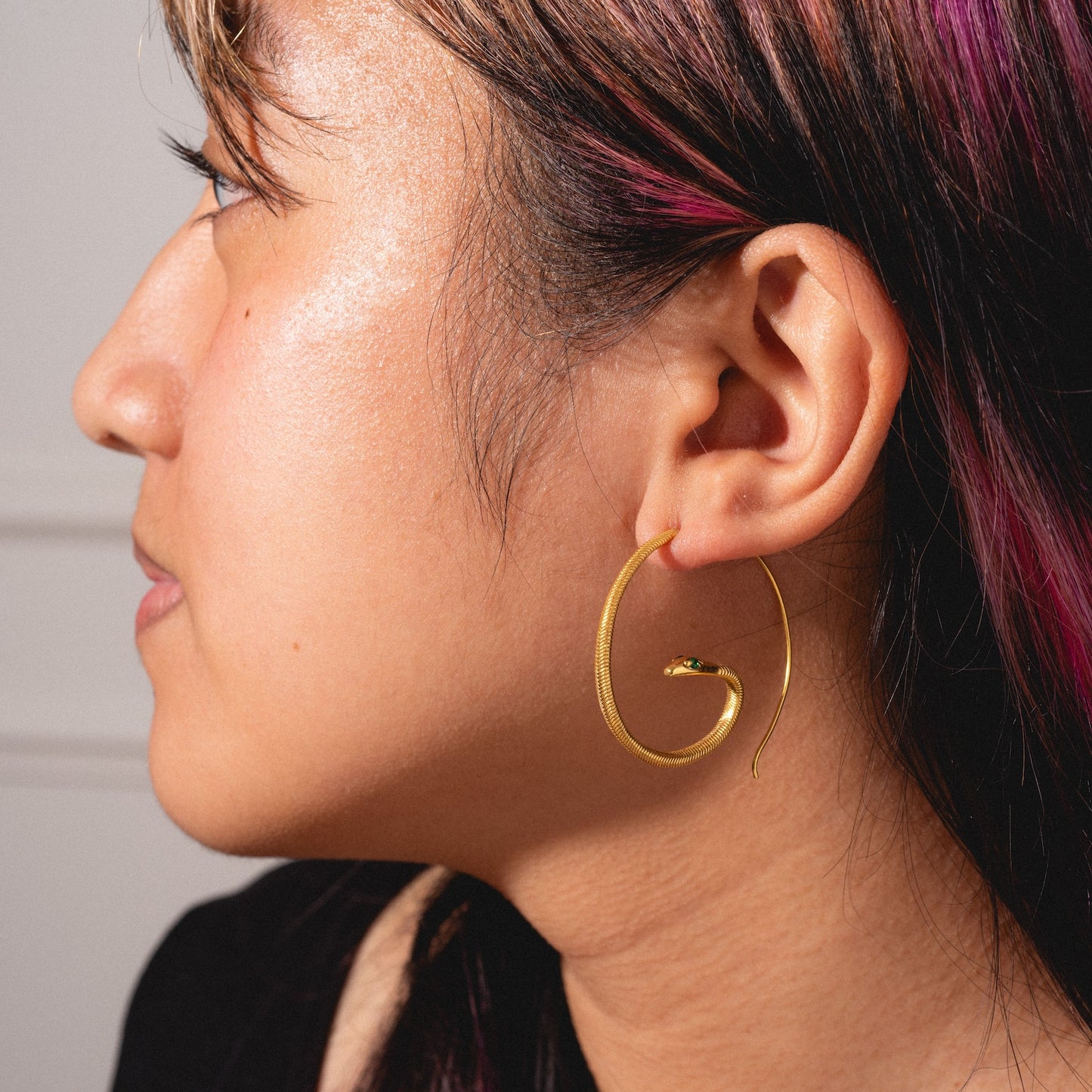 A person with dark hair and pink highlights wears a Mini Snake Hoop earring in their left ear. The close-up profile photo highlights the unique design, showing their ear, jawline, and part of their face.