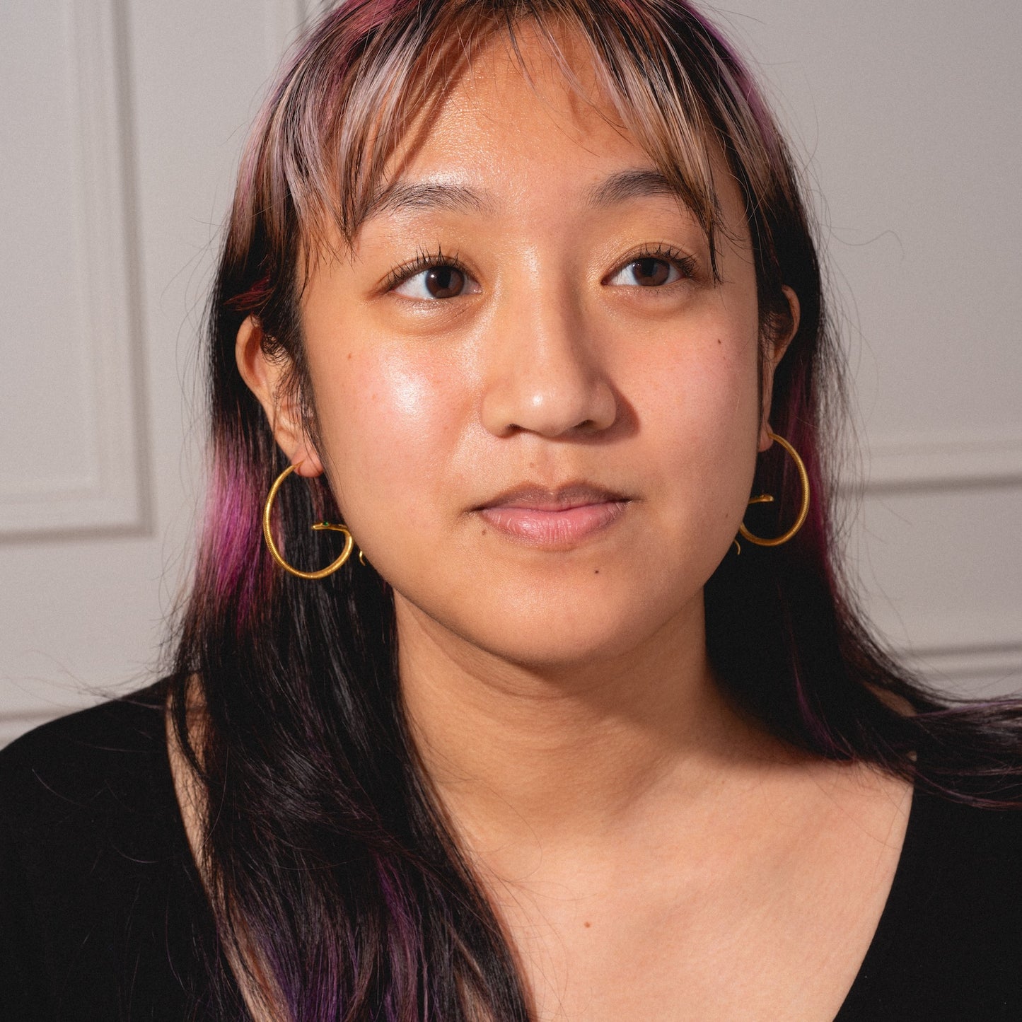 A woman with long dark hair and pink highlights wears Mini Snake Hoops and a black top, gazing slightly to the side with a neutral expression against a light-colored background.