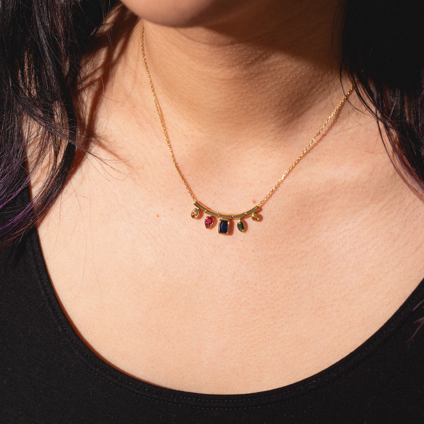 A person wearing the Technicolor Gem Drop Necklace, featuring five colorful gemstone charms—red, green, and blue—against bare skin and a black top.