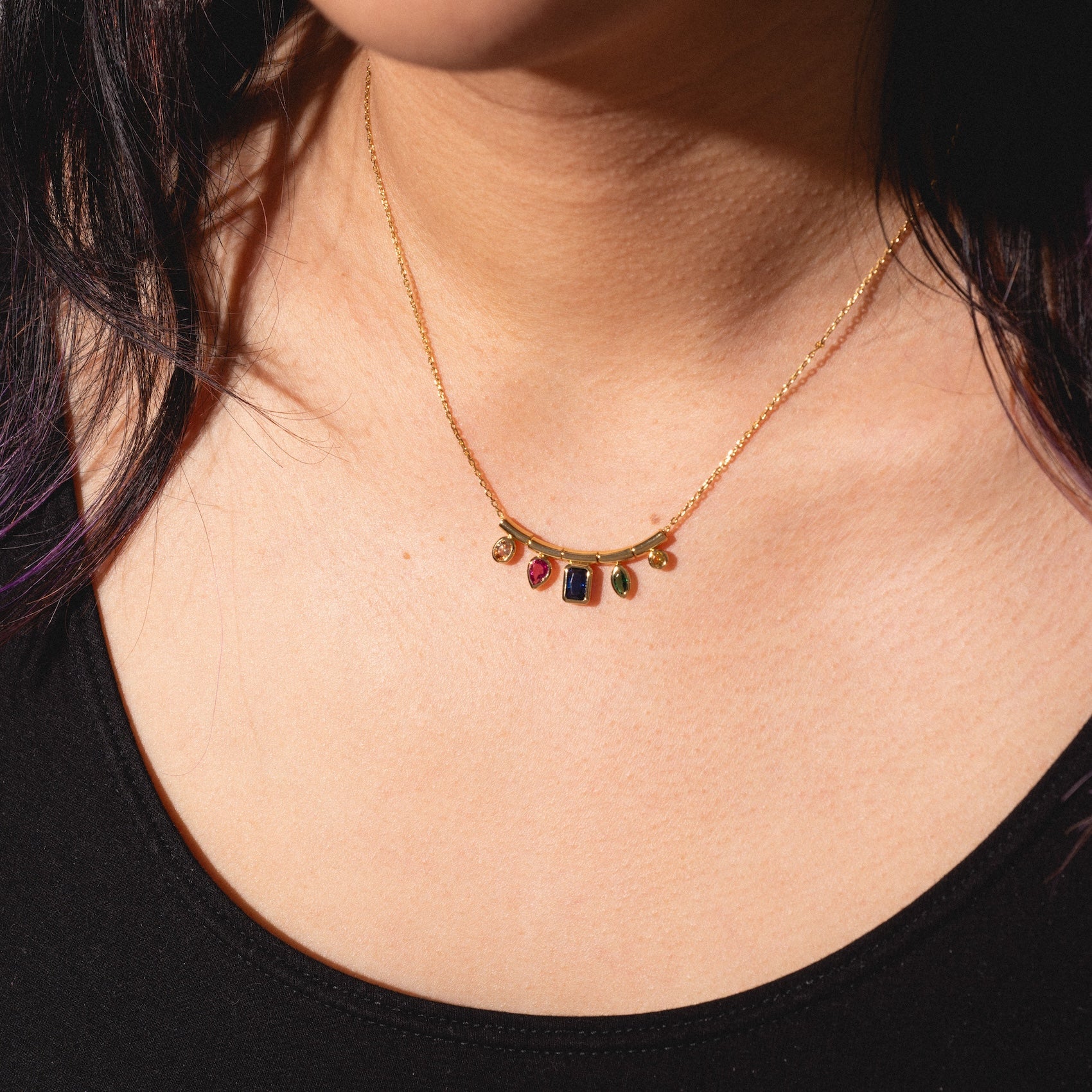 A person wearing the Technicolor Gem Drop Necklace, featuring five colorful gemstone charms—red, green, and blue—against bare skin and a black top.