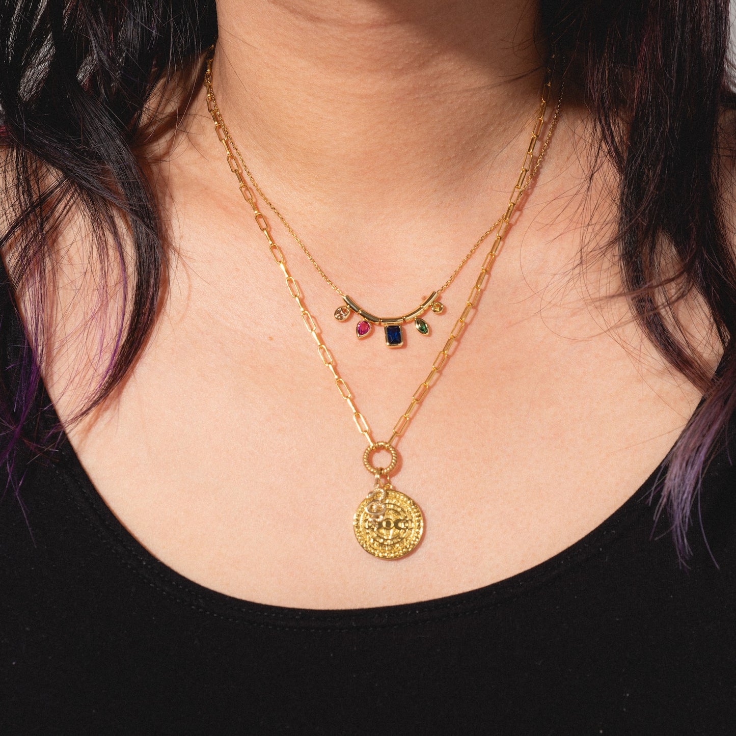A woman wears two Charms by Jurate JLA yellow gold plated necklaces: one with a large round medallion pendant and another featuring small colorful gemstone charms, layered over a black top.