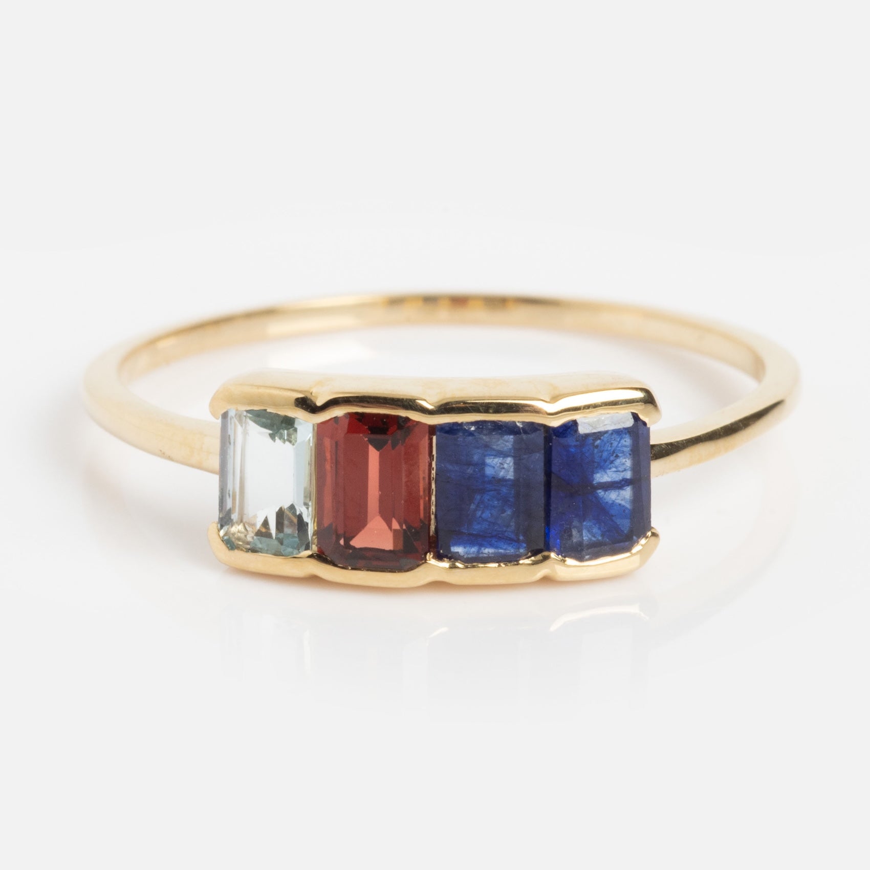 A solid gold ring featuring baguette-cut aquamarine, sapphire, and garnet gemstones set side by side in a simple band. Shown in sample size 9 against a white background.