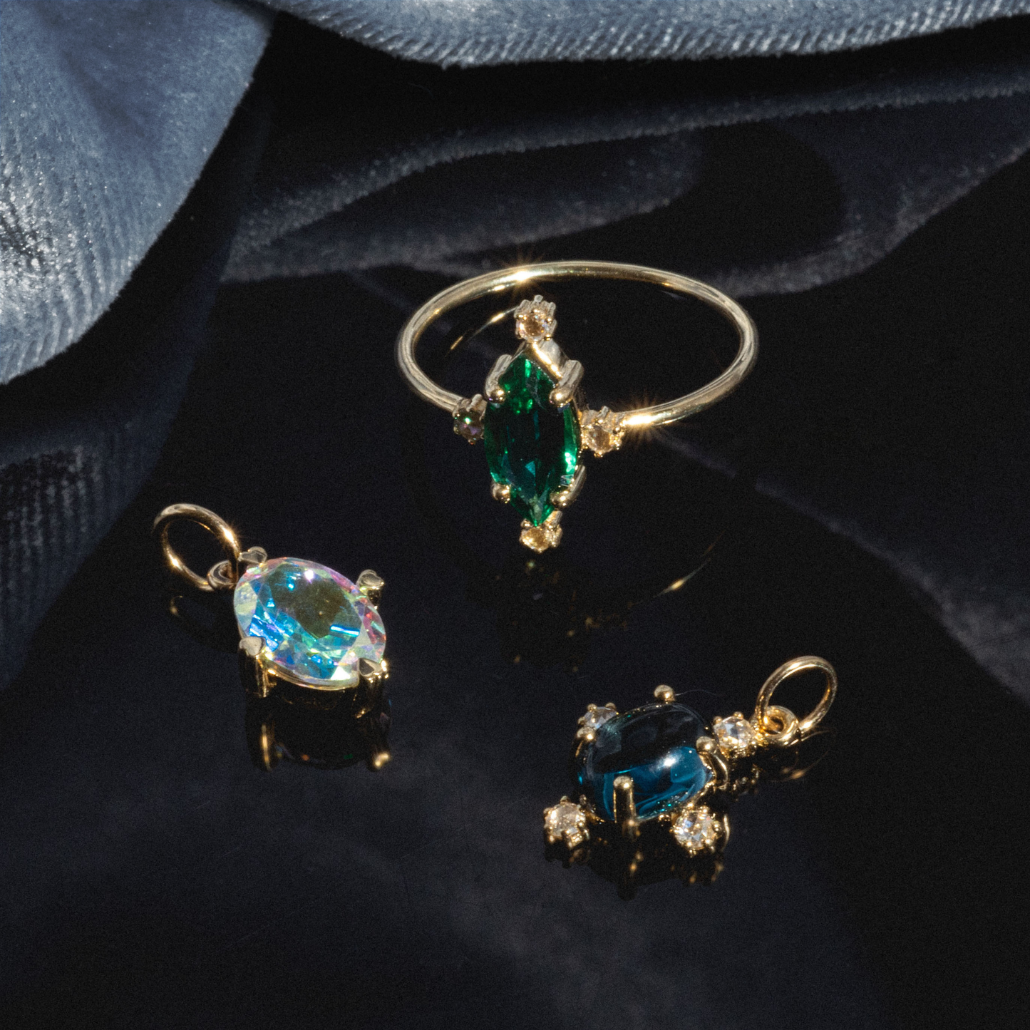 A One of a Kind 14k Marquis Lab Emerald and Diamond Ring is displayed on a shiny black surface, accompanied by two gold pendants—one with an iridescent oval gem and one with a dark blue oval stone—against a blue fabric background.