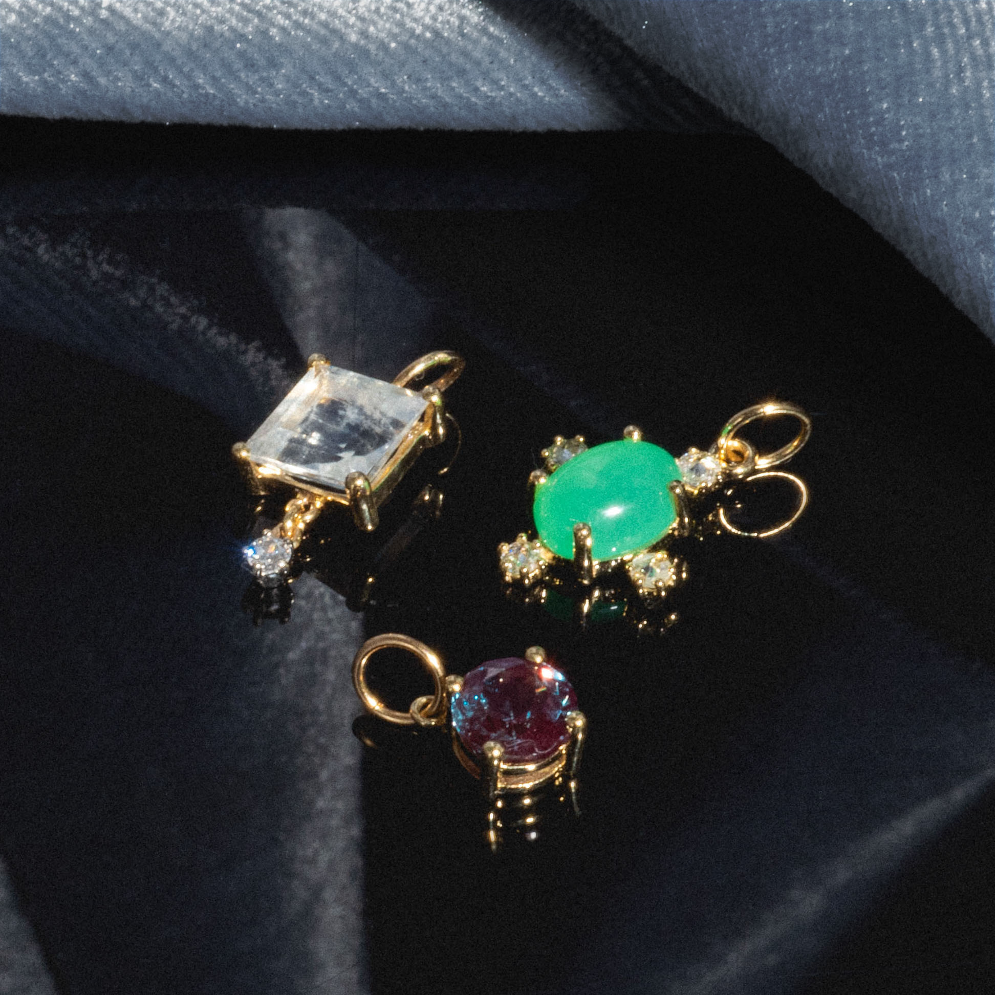 The One of a Kind 14k Round Lab Alexandrite Charm, along with two other gold pendants—a square clear gem and an oval green stone with gems—rests on a glossy dark surface against soft blue fabric.