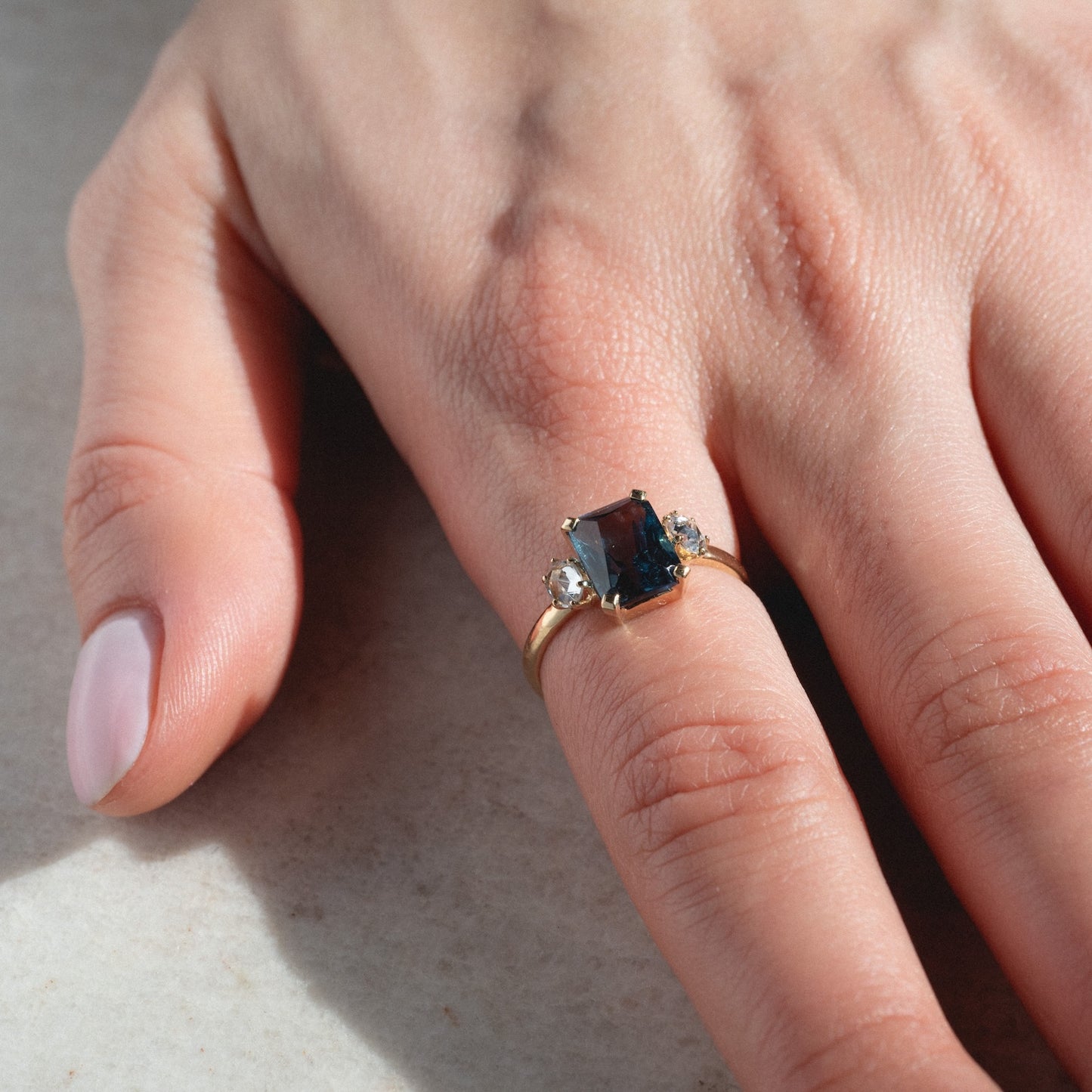 A hand with light pink polished nails wears the One of a Kind 14k Vintage Inspired London Blue Topaz and White Sapphire Ring, featuring a rectangular blue topaz flanked by white sapphires, resting on a light-colored surface.