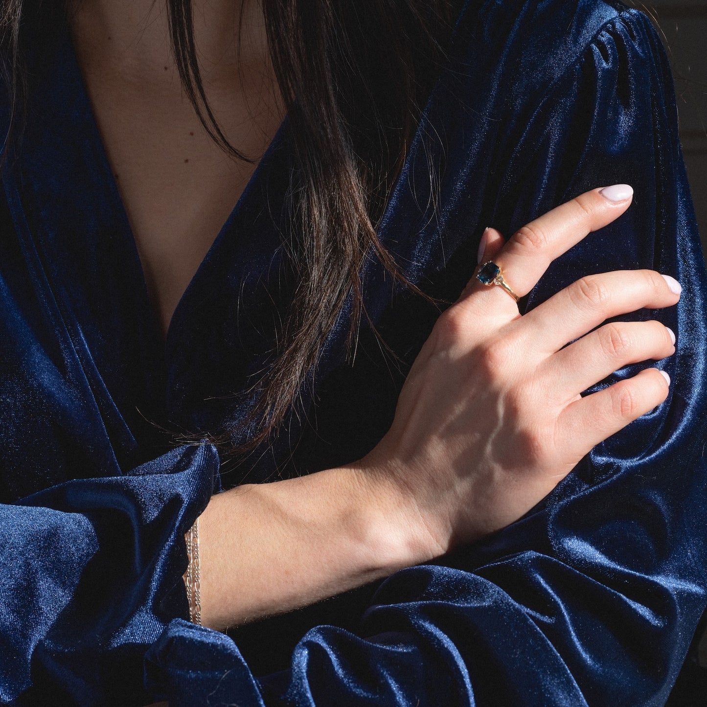 A person in a deep blue velvet blouse rests their hand on their arm, showcasing the One of a Kind 14k Vintage Inspired London Blue Topaz and White Sapphire Ring. Their face is partially out of frame, with dark hair falling over one shoulder.