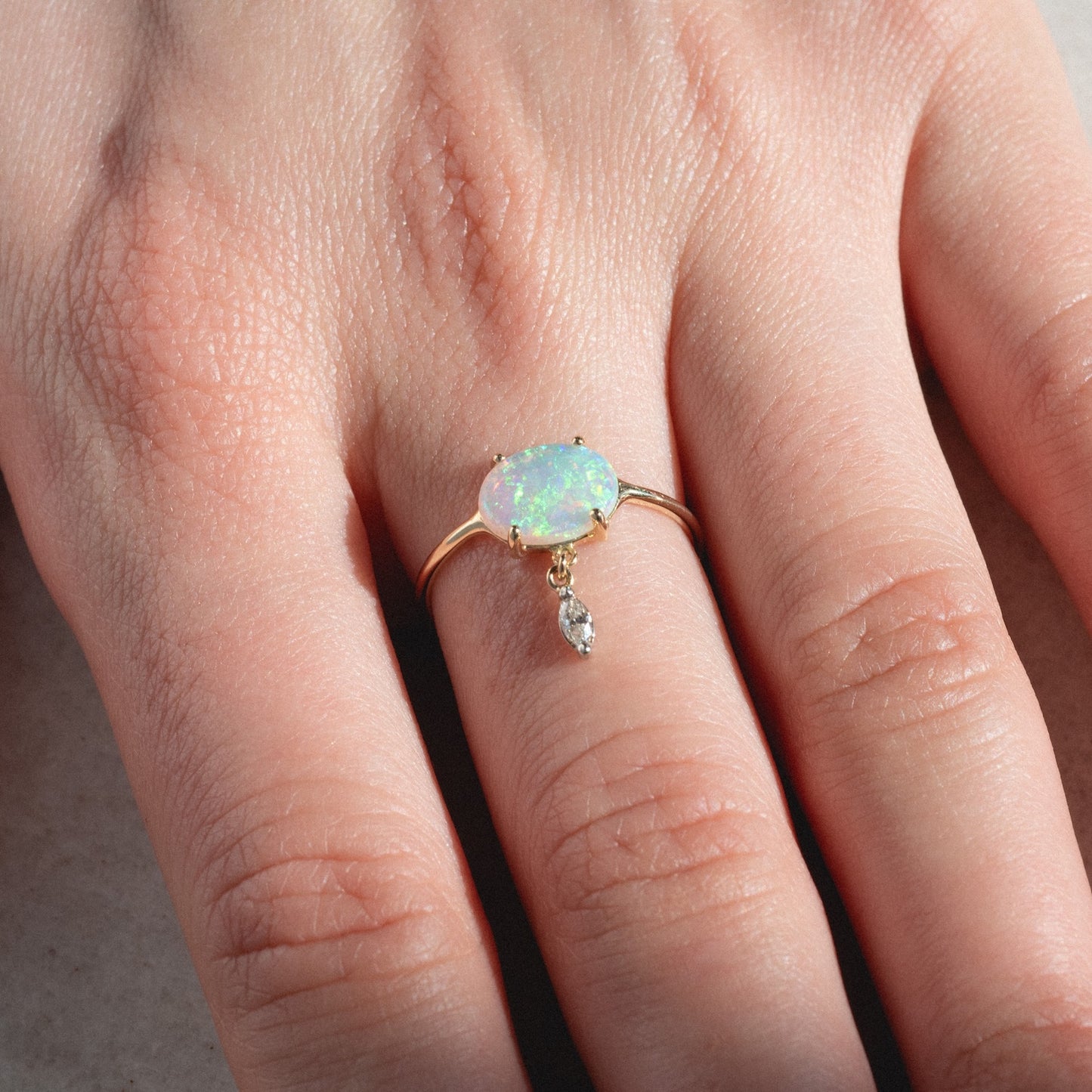 A hand wears the One of a Kind 14k Opal with Diamond Dangle Ring, featuring an oval opal and a dangling marquise-shaped diamond charm, displayed against a neutral background.