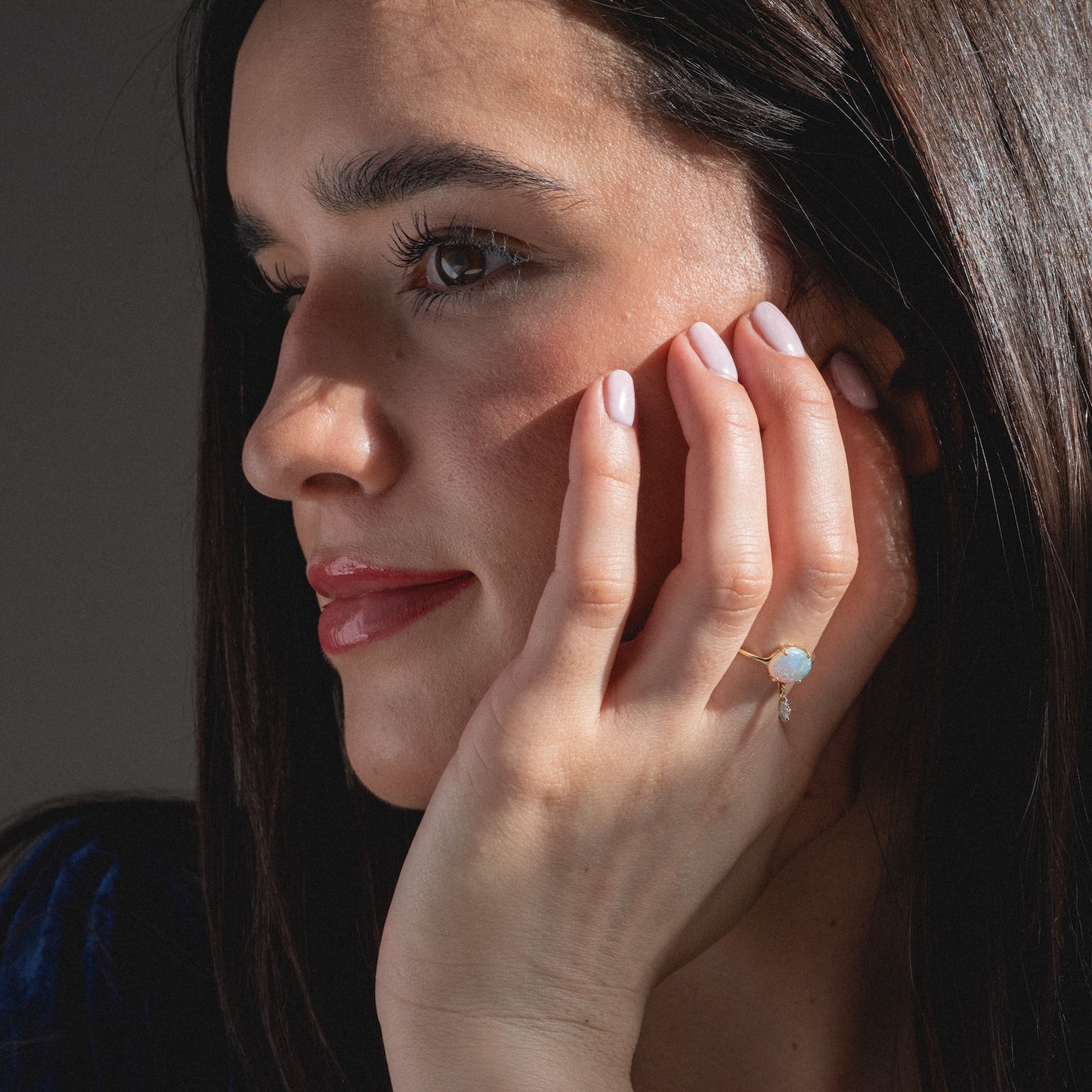 A woman with long brown hair rests her chin on her hand, showcasing the One of a Kind 14k Opal with Diamond Dangle Ring. With natural makeup and glossy lips, she gazes thoughtfully to the side.