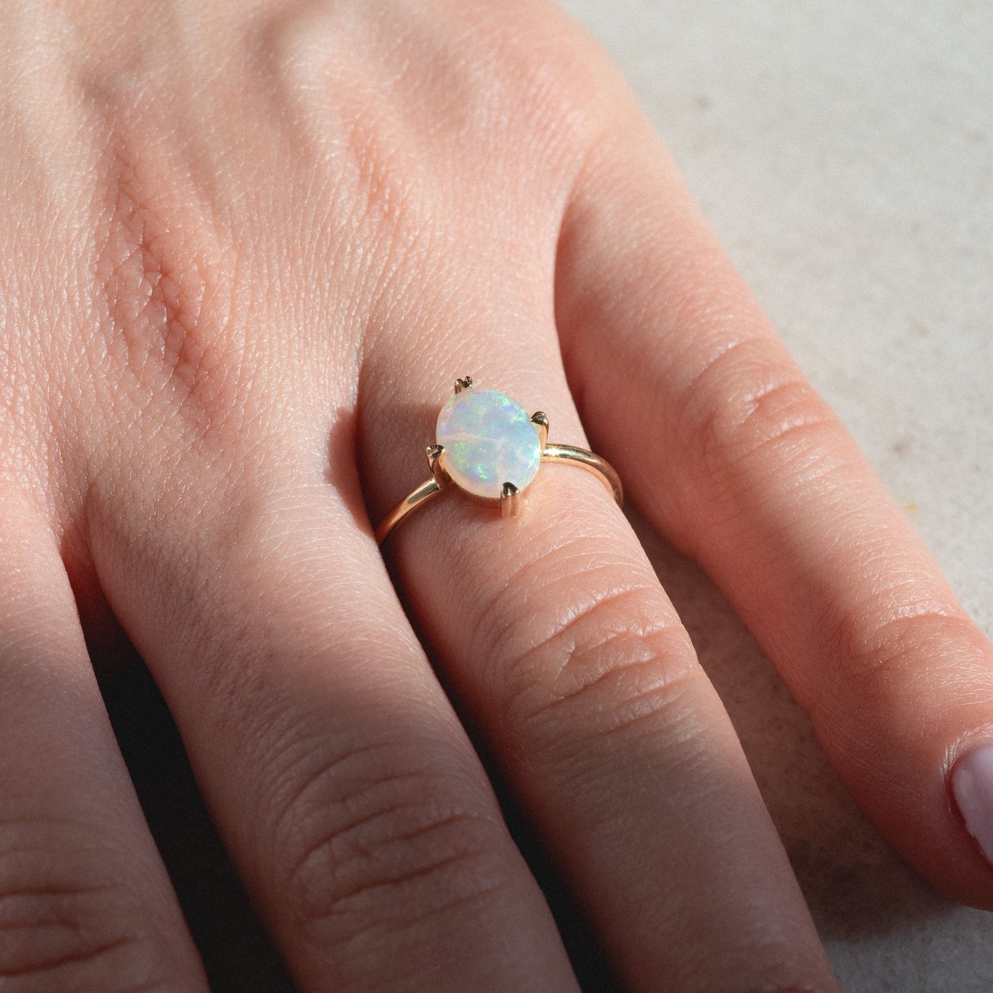 A close-up of a hand wearing the One of a Kind 14k Heart Prong Opal Ring, featuring an oval opal gemstone on the ring finger, resting on a light, textured surface.
