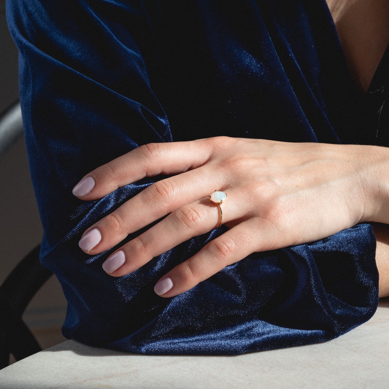 A hand with light pink nails rests on marble, wearing the One of a Kind 14k Heart Prong Opal Ring and dressed in deep blue velvet.