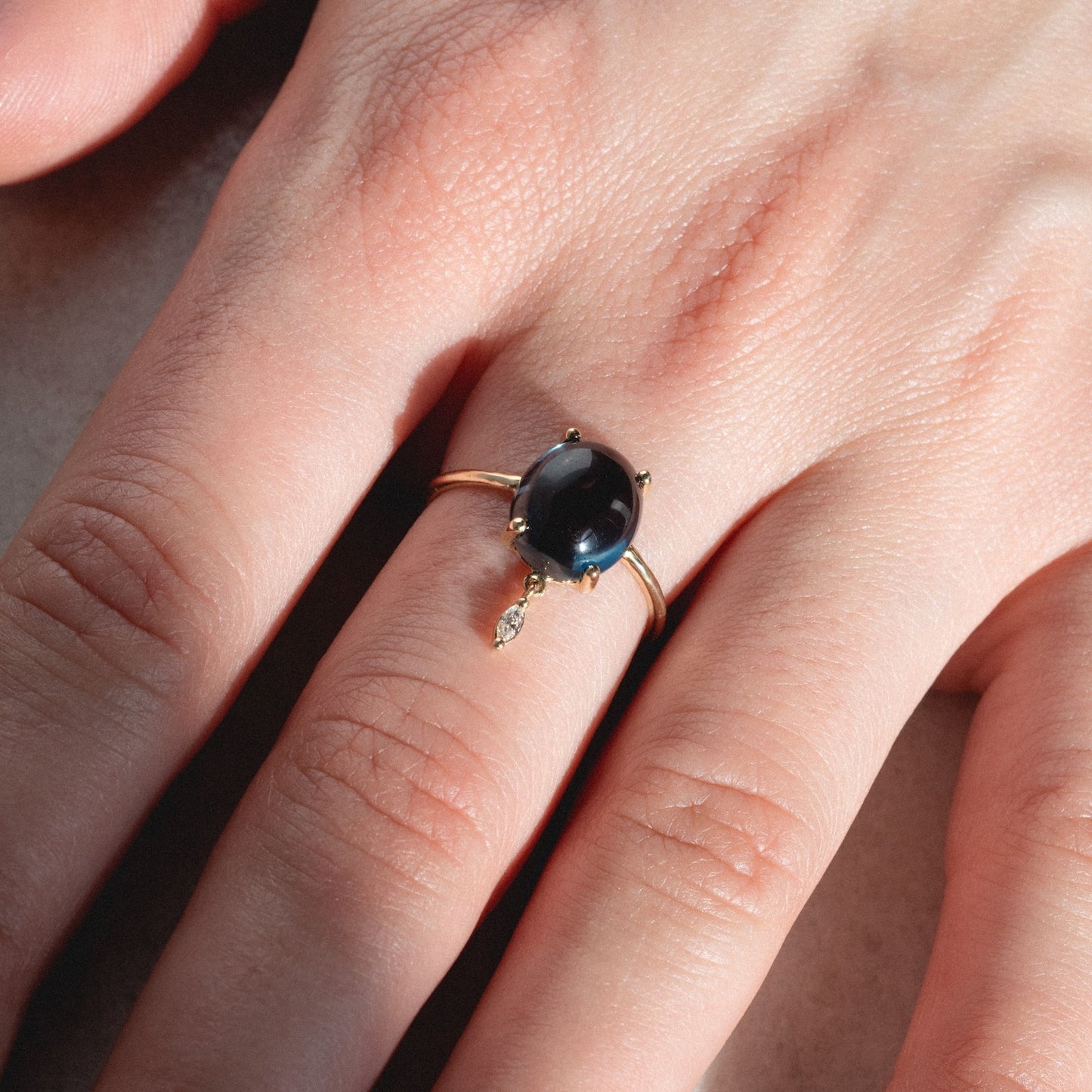 A close-up of a hand wearing the One of a Kind 14k London Blue Topaz with Diamond Dangle Ring, highlighting the dark oval gemstone and sparkling diamond charm against natural skin.