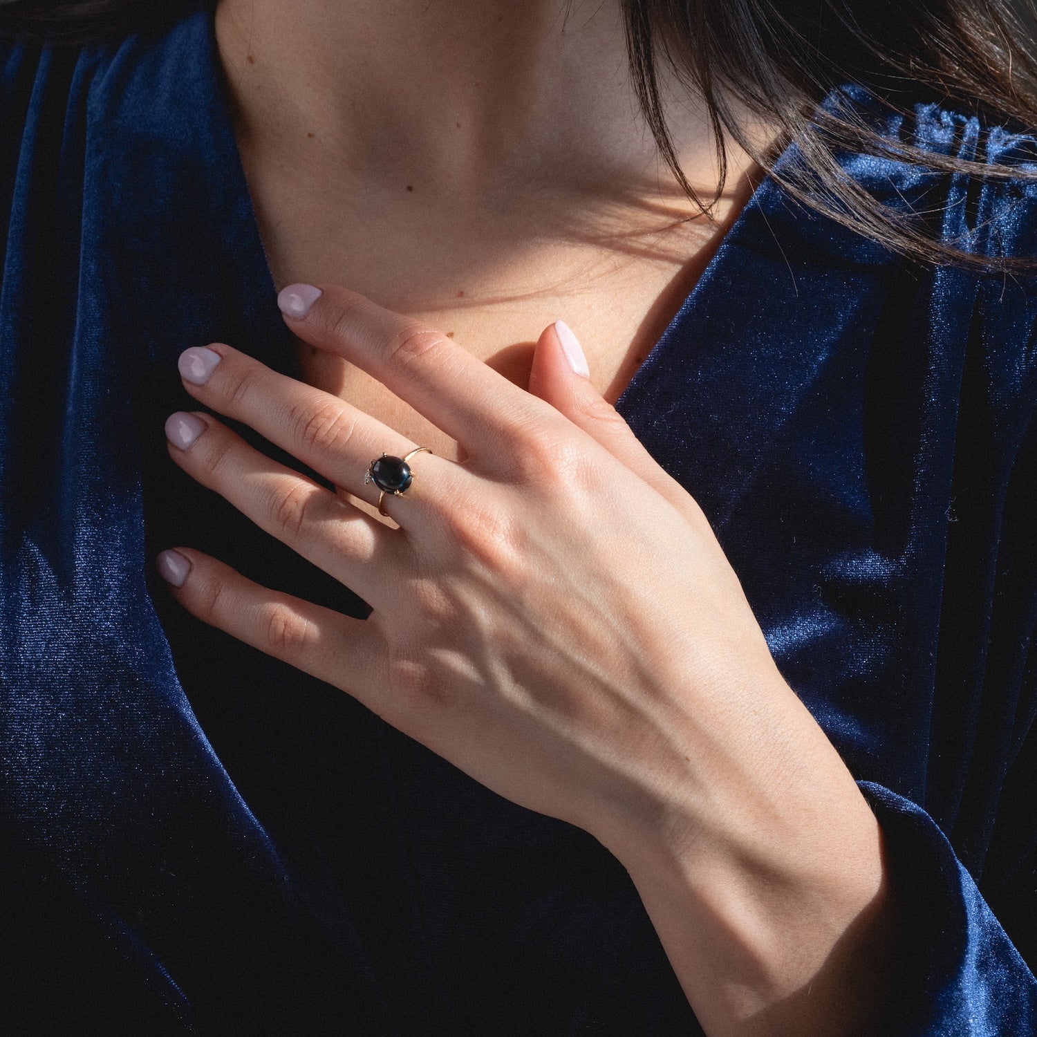 A woman in a deep blue velvet top rests her hand on her chest, showing off the One of a Kind 14k London Blue Topaz with Diamond Dangle Ring on her finger.
