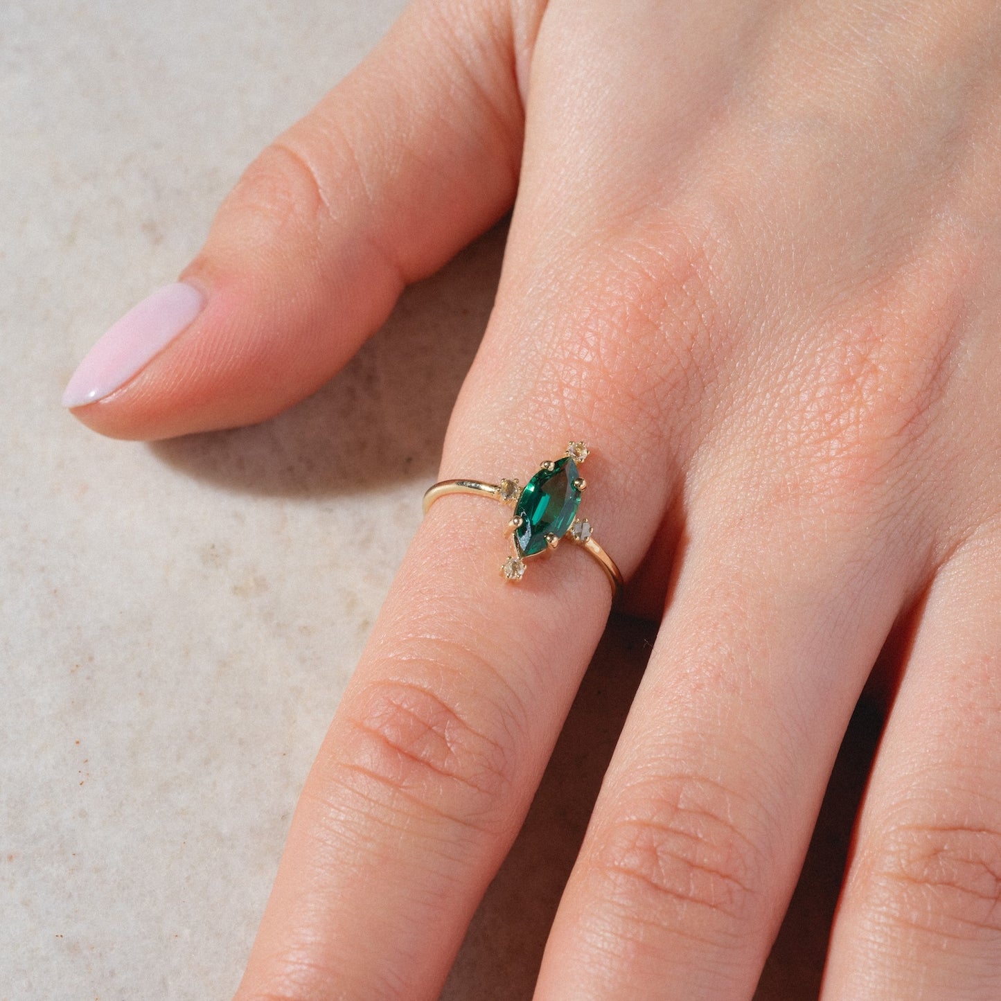 A hand with light skin wears the One of a Kind 14k Marquis Lab Emerald and Diamond Ring, featuring an oval green gemstone and small clear stones on the ring finger. The nails are neatly manicured and painted pale pink.