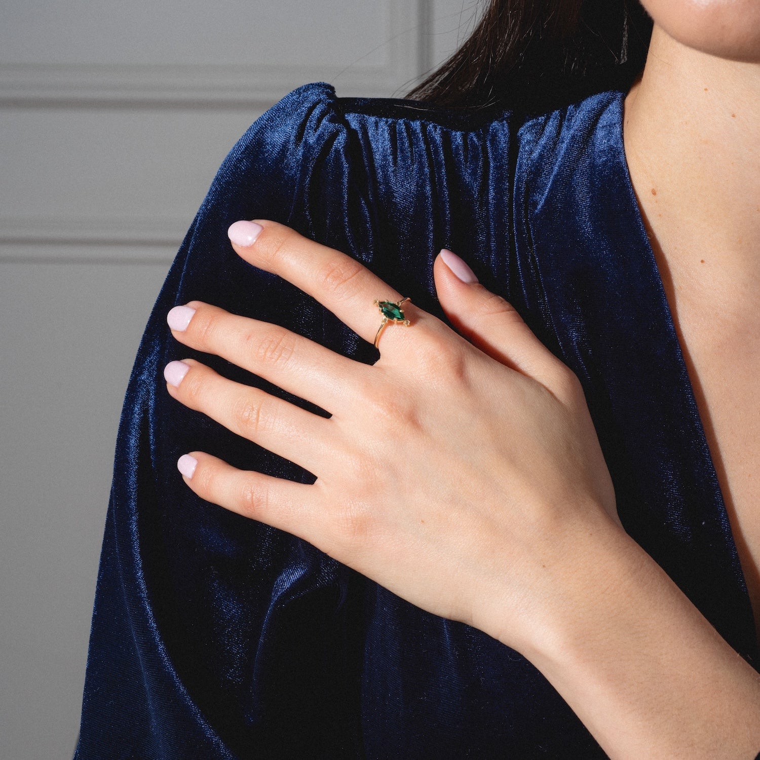 A woman in a deep blue velvet dress rests her hand on her chest, showing off the One of a Kind 14k Marquis Lab Emerald and Diamond Ring on her finger. Her nails are manicured with light pink polish.