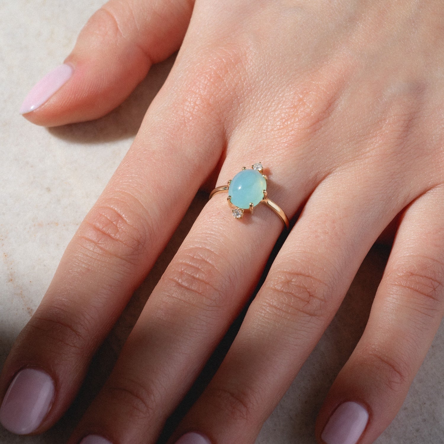 A hand with pale pink polished nails wears the One of a Kind 14k Peruvian Opal and Diamond Ring, featuring a light blue oval opal surrounded by diamonds, resting on a light surface.
