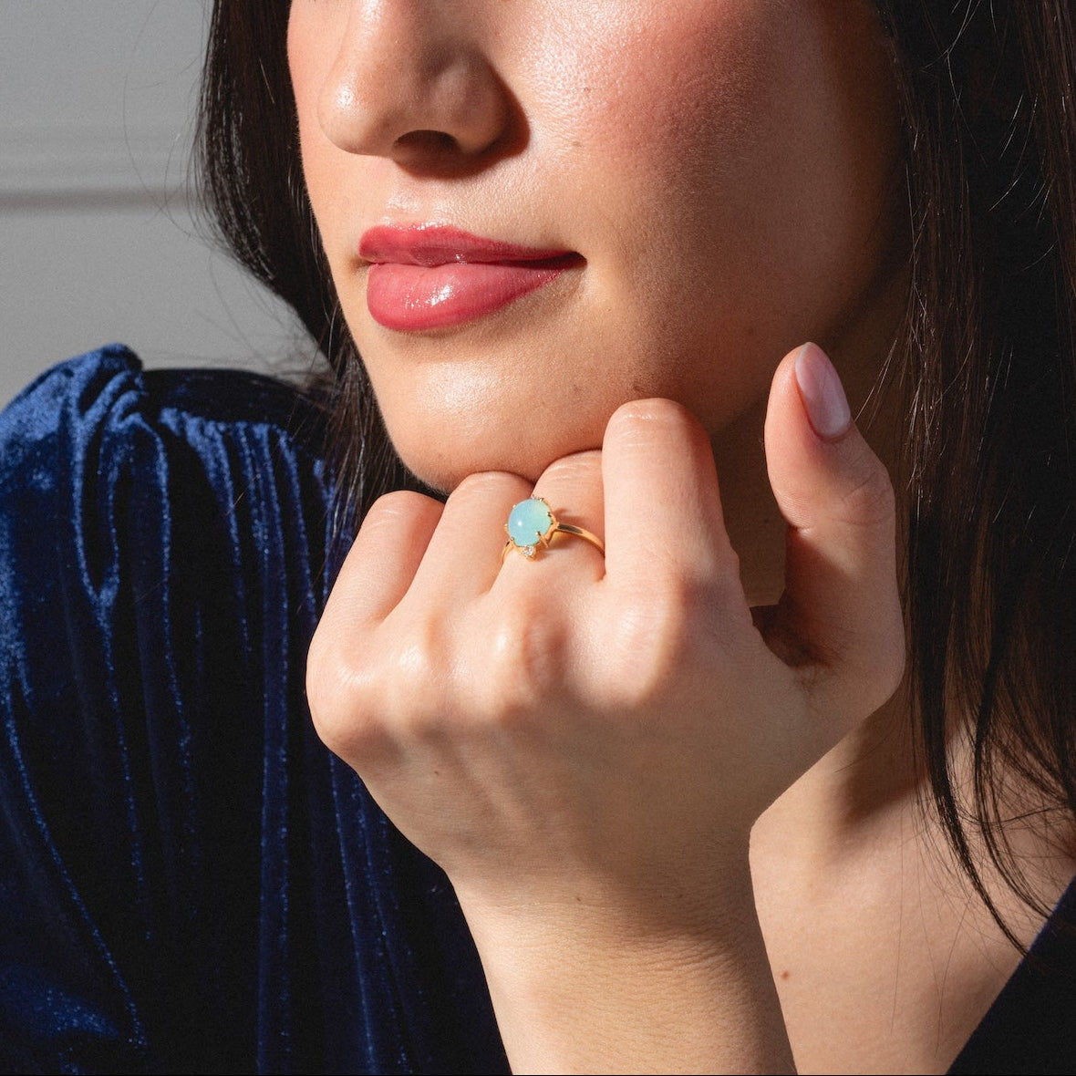 A woman with long dark hair rests her chin on her hand, showcasing the One of a Kind 14k Peruvian Opal and Diamond Ring. She wears a dark blue velvet top and gazes thoughtfully to the side.