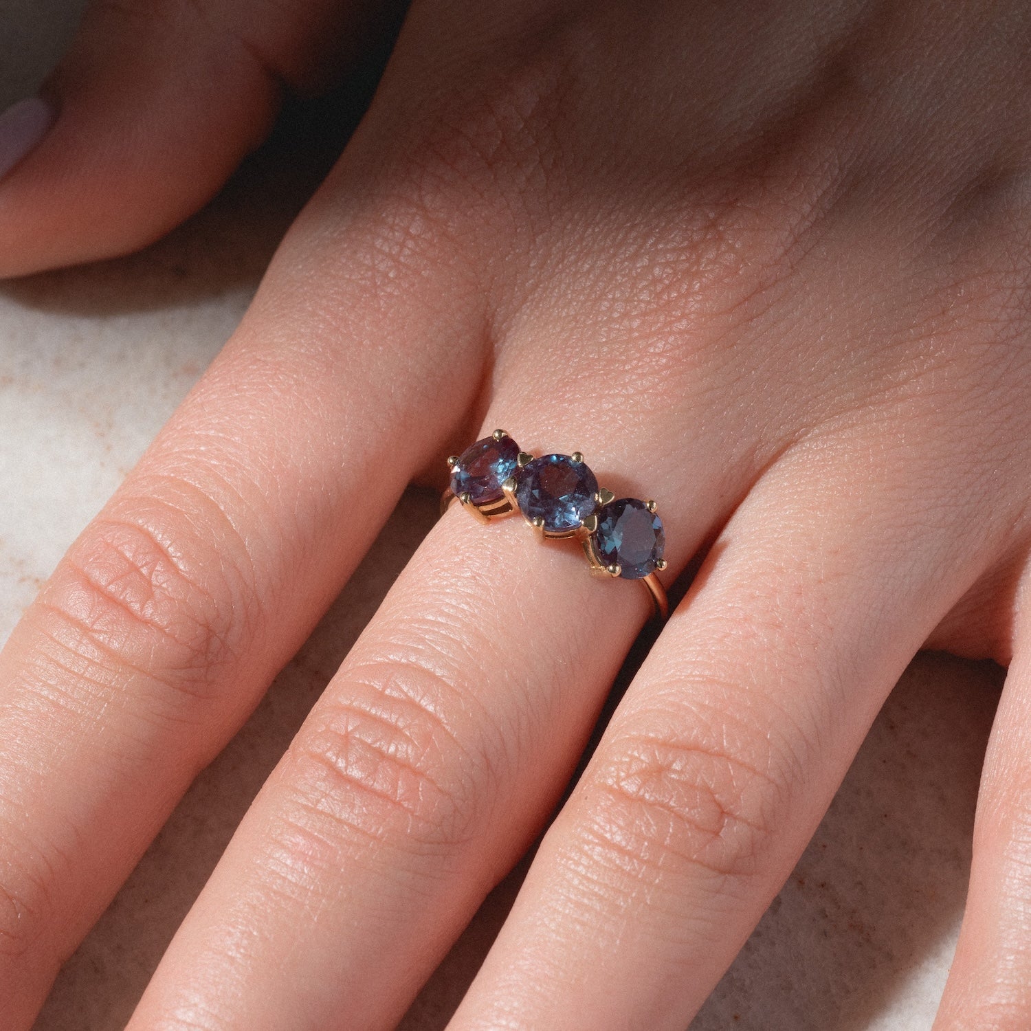 A close-up of a hand wearing the One of a Kind 14k Heart Prong Lab Alexandrite Trio Ring, featuring three oval-shaped lab alexandrite gemstones set side by side on the ring finger, resting on a light surface.