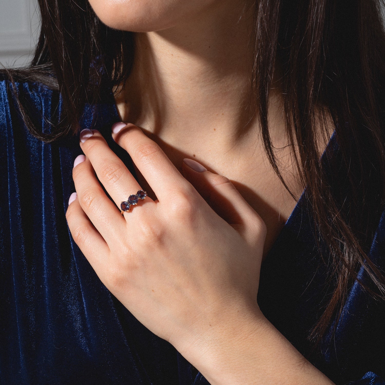 A woman wearing a deep blue velvet top displays her manicured hand adorned with the One of a Kind 14k Heart Prong Lab Alexandrite Trio Ring, featuring three dark gemstones set in a row. Her face is partially visible.