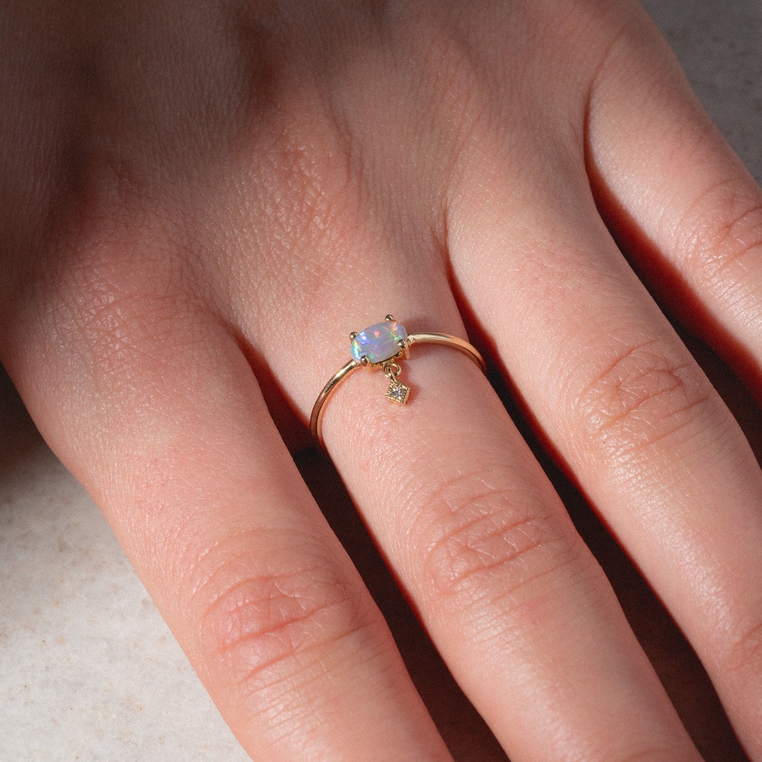 A hand displays the One of a Kind 14k Sweetest Opal with Diamond Dangle Ring—featuring a delicate opal and tiny diamond charm—on the middle finger against a neutral background.