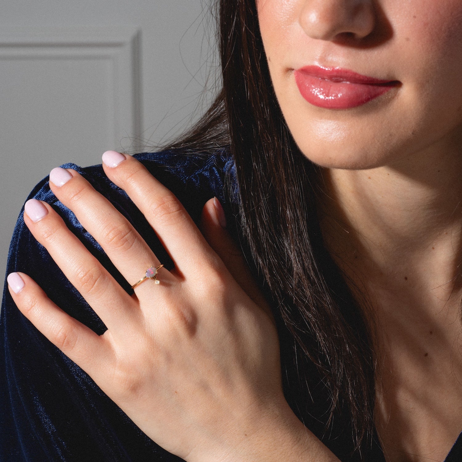 A woman in a dark blue velvet top rests her hand on her shoulder, showing off the One of a Kind 14k Sweetest Opal with Diamond Dangle Ring. Her nails are light pink and her lips have a glossy pink finish.