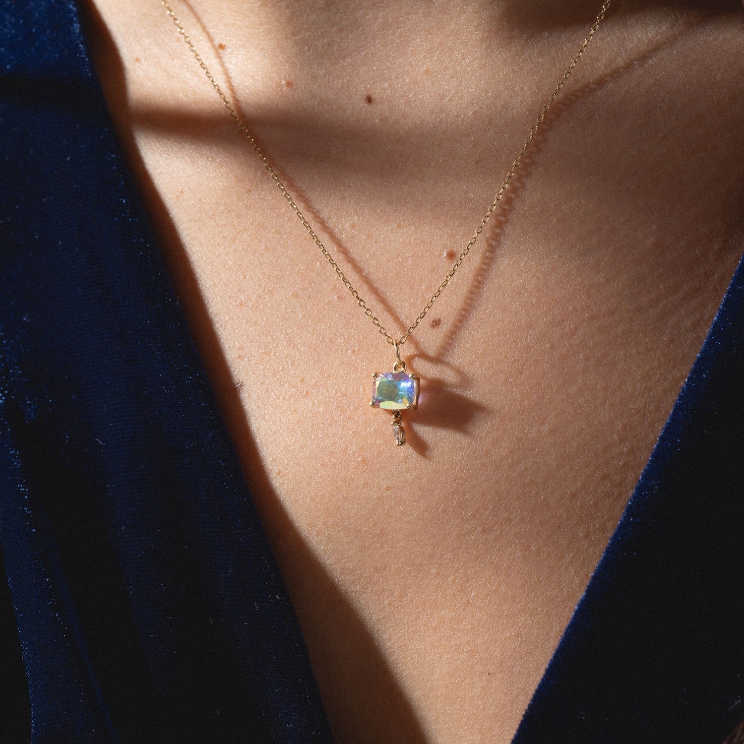 Close-up of a person wearing the One of a Kind 14k Pillow Cut Angel Aura with Diamond Dangle Charm necklace. The square pendant dazzles with multi-colored reflections against bare skin and a dark blue V-neck garment.