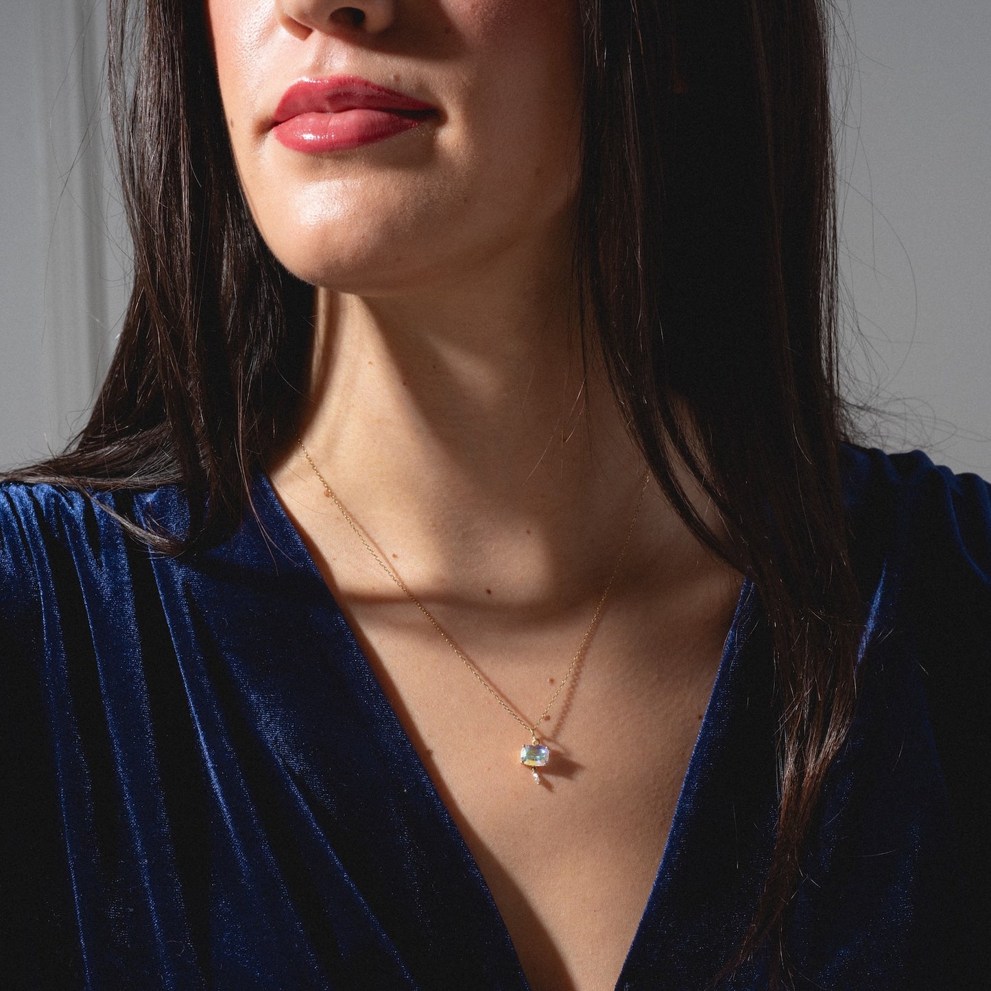 A woman wears a deep blue velvet top and the One of a Kind 14k Pillow Cut Angel Aura with Diamond Dangle Charm necklace. She has loose dark hair, natural makeup, and pink lipstick, shown from the shoulders up.