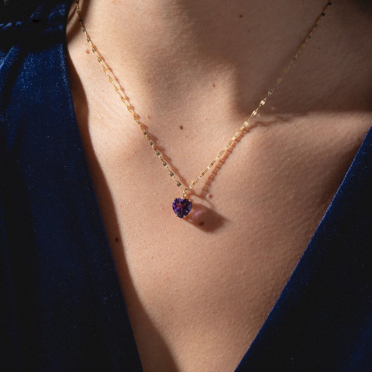A close-up of a person wearing the One of a Kind 14k Amethyst Heart Charm necklace, featuring a delicate gold chain and purple gemstone pendant against bare skin and a deep blue velvet V-neck top.