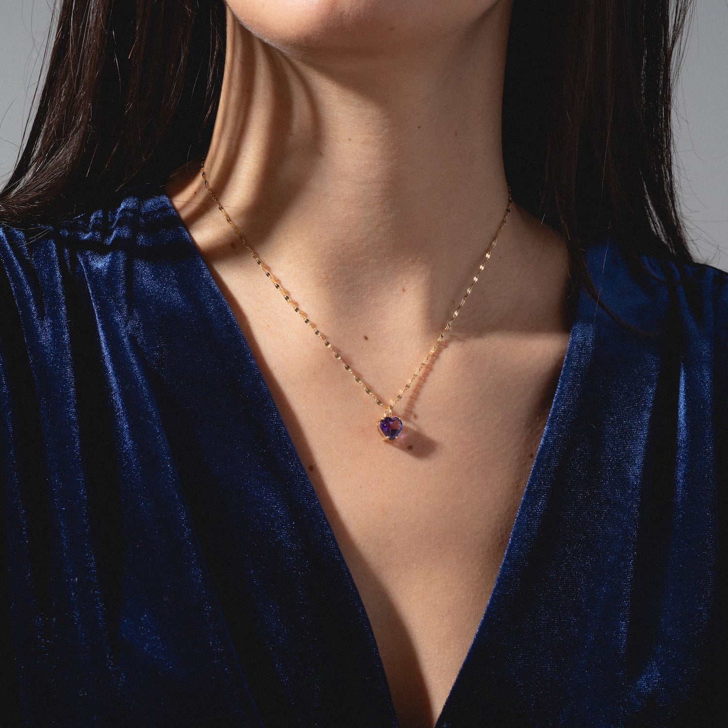 A woman wearing a deep blue velvet top and a delicate gold necklace featuring the One of a Kind 14k Amethyst Heart Charm, shown from the shoulders to just above the lips.