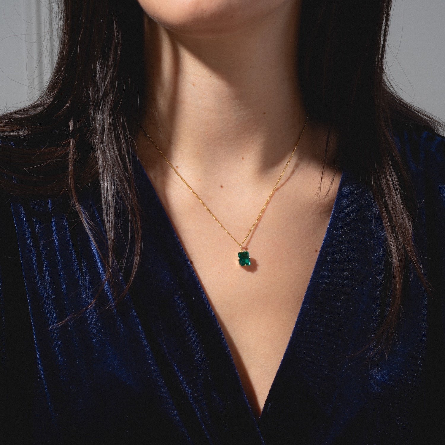 A woman in a deep blue velvet top highlights the One of a Kind 14k Lab Emerald Charm with Hand Engraved Details—a delicate gold necklace featuring a vibrant green gemstone pendant against her bare neck and chest.