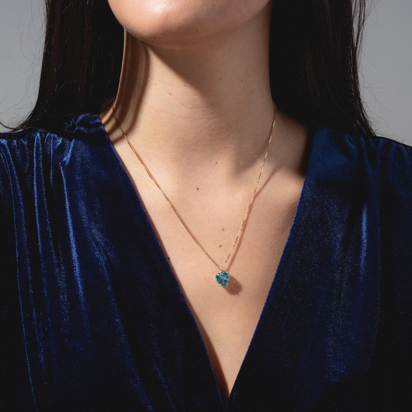 A woman wears a deep blue velvet top and the One of a Kind 14k Blue Topaz Heart Charm necklace, shown from the shoulders up against a neutral background.
