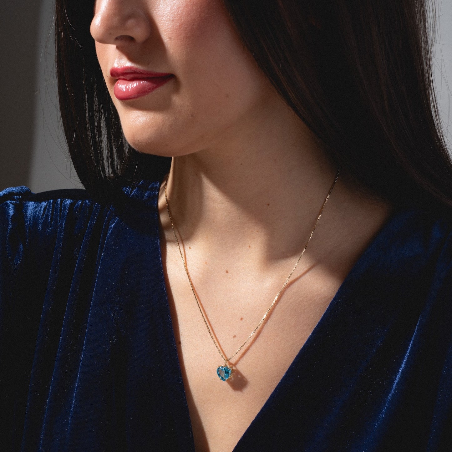 A woman wears a deep blue velvet top and a thin gold necklace featuring the One of a Kind 14k Blue Topaz Heart Charm, shown from the shoulders up with her face partly visible and lips painted red.