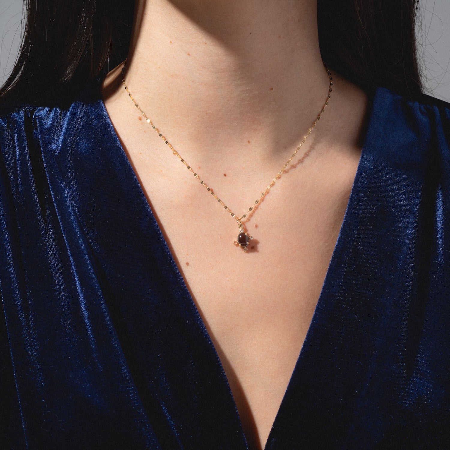 A person wears a deep blue velvet V-neck top and the One of a Kind 14k London Blue Topaz and Diamond Charm necklace, shown from shoulders to chest against a neutral background.