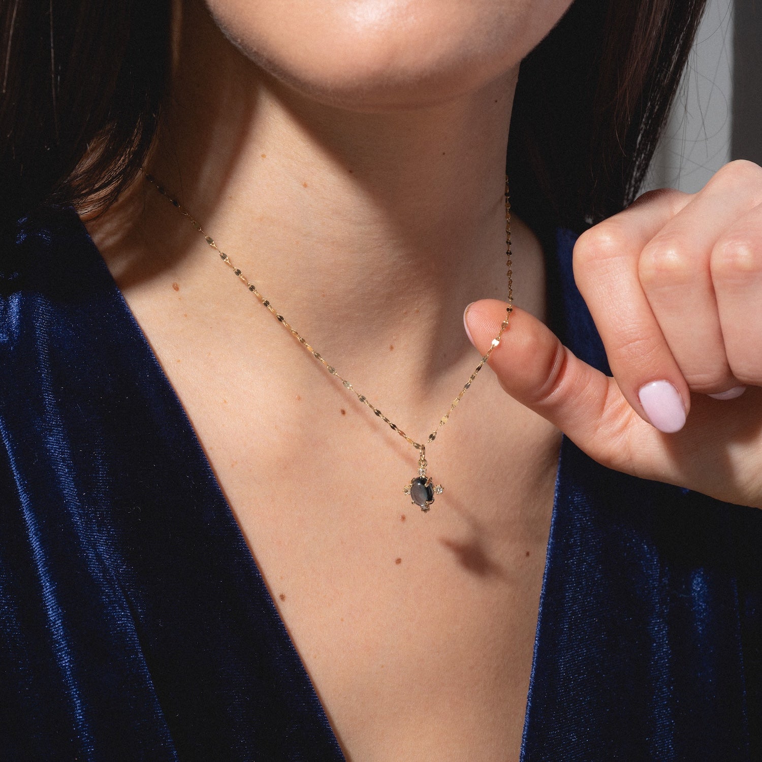 A woman in a deep blue velvet top holds the One of a Kind 14k London Blue Topaz and Diamond Charm necklace near her neck. Her light pink nails are visible as the photo focuses on her neck, collarbone, and hand.