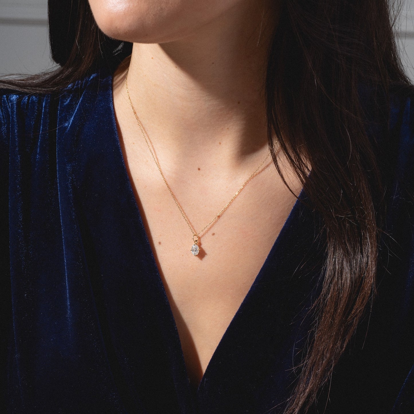 A woman wears a deep blue velvet top and the One of a Kind 14k Oval Moissanite Charm with Hand Engraved Details, a delicate gold necklace, shown from shoulders to chin.