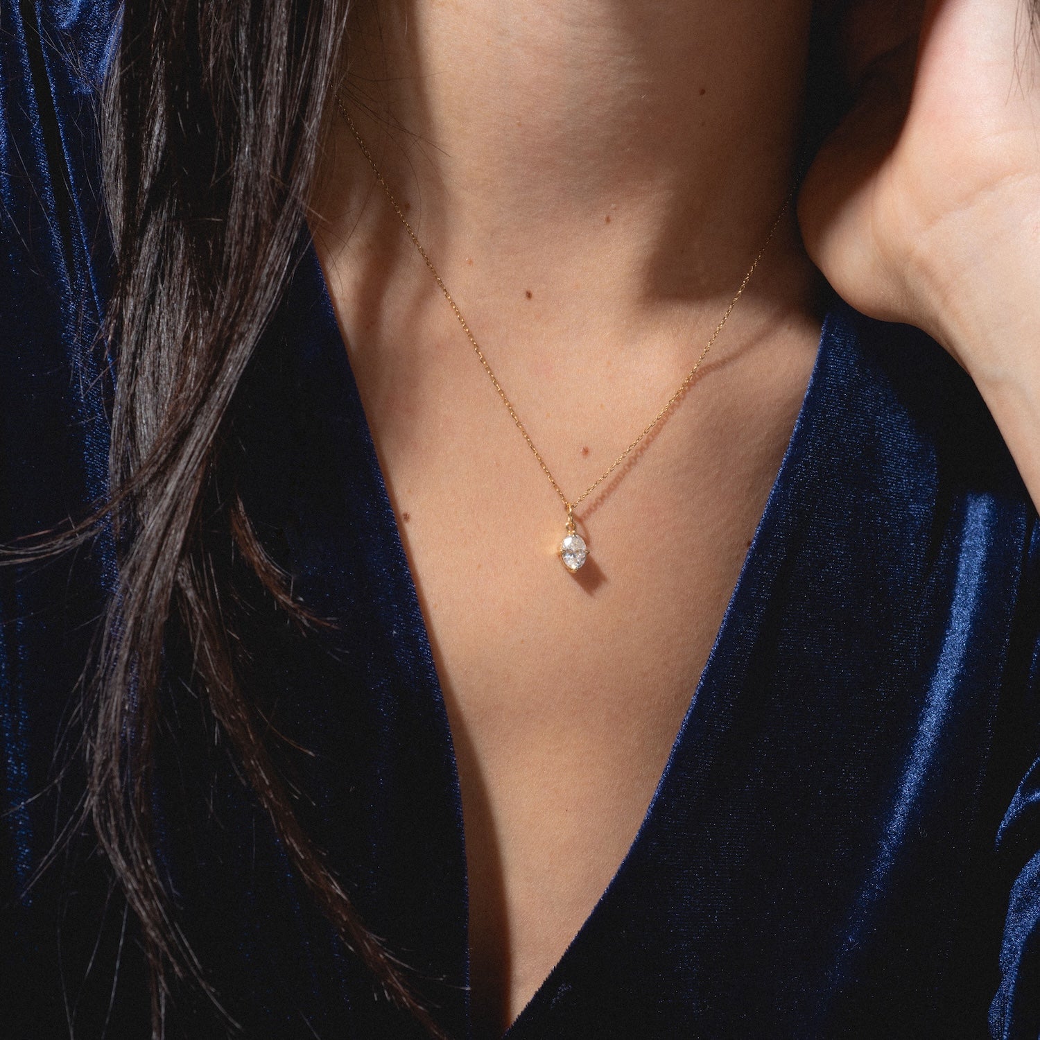 A close-up of a woman in a dark blue velvet top wearing the One of a Kind 14k Oval Moissanite Charm with Hand Engraved Details. Her hand is near her face, and her dark hair falls over her shoulder.