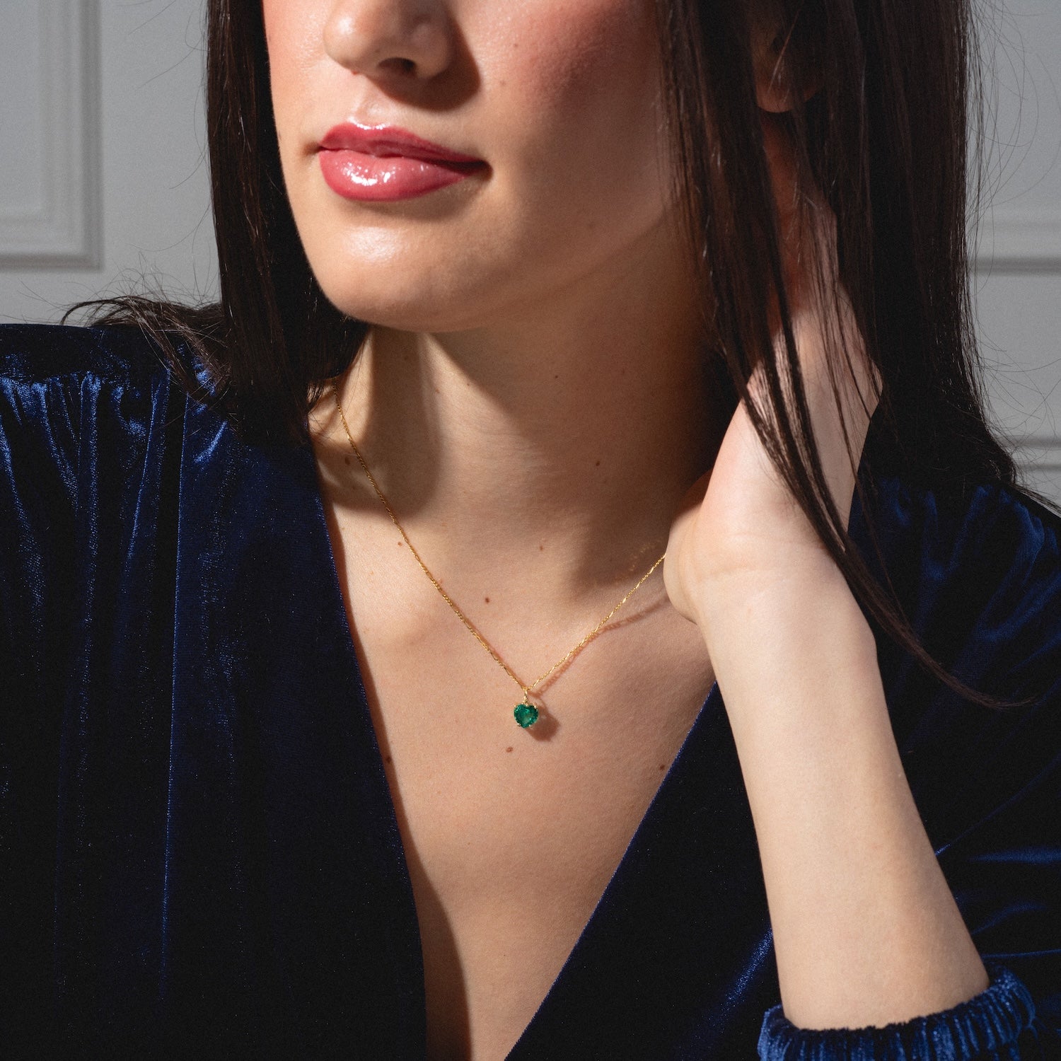 A woman in a deep blue velvet top wears the One of a Kind 14k Lab Emerald Heart Charm, a delicate gold necklace with a small green heart pendant. She touches her neck with one hand; only her lips, lower face, and upper torso are visible.