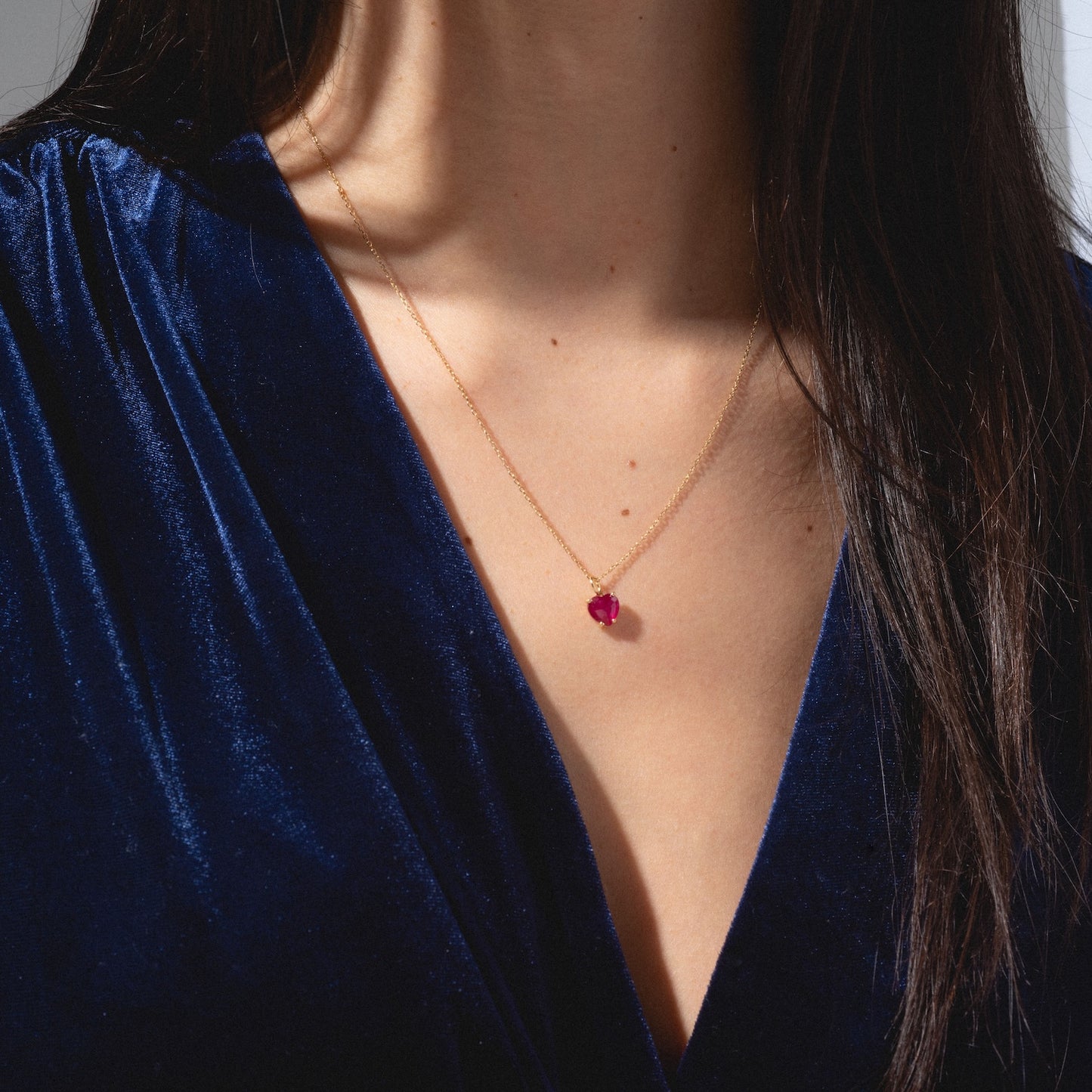 A woman wearing a deep blue velvet dress and a One of a Kind 14k Ruby Heart Charm necklace. Only her neck, upper chest, and part of her dark hair are visible.