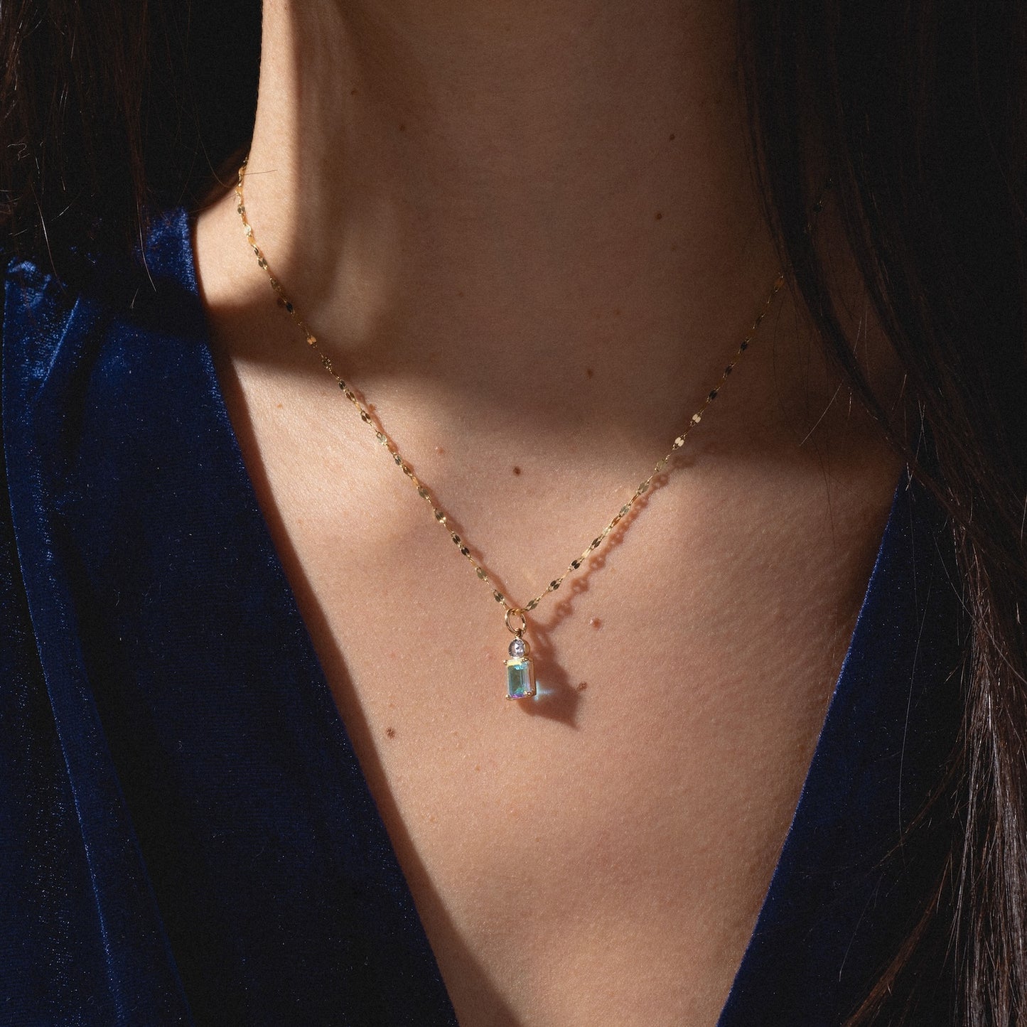 A close-up of a person wearing the One of a Kind 14k Angel Aura and Diamond Charm necklace, featuring a small pendant with a light blue gemstone, against a deep blue velvet top. The focus is on the unique necklace and charm.