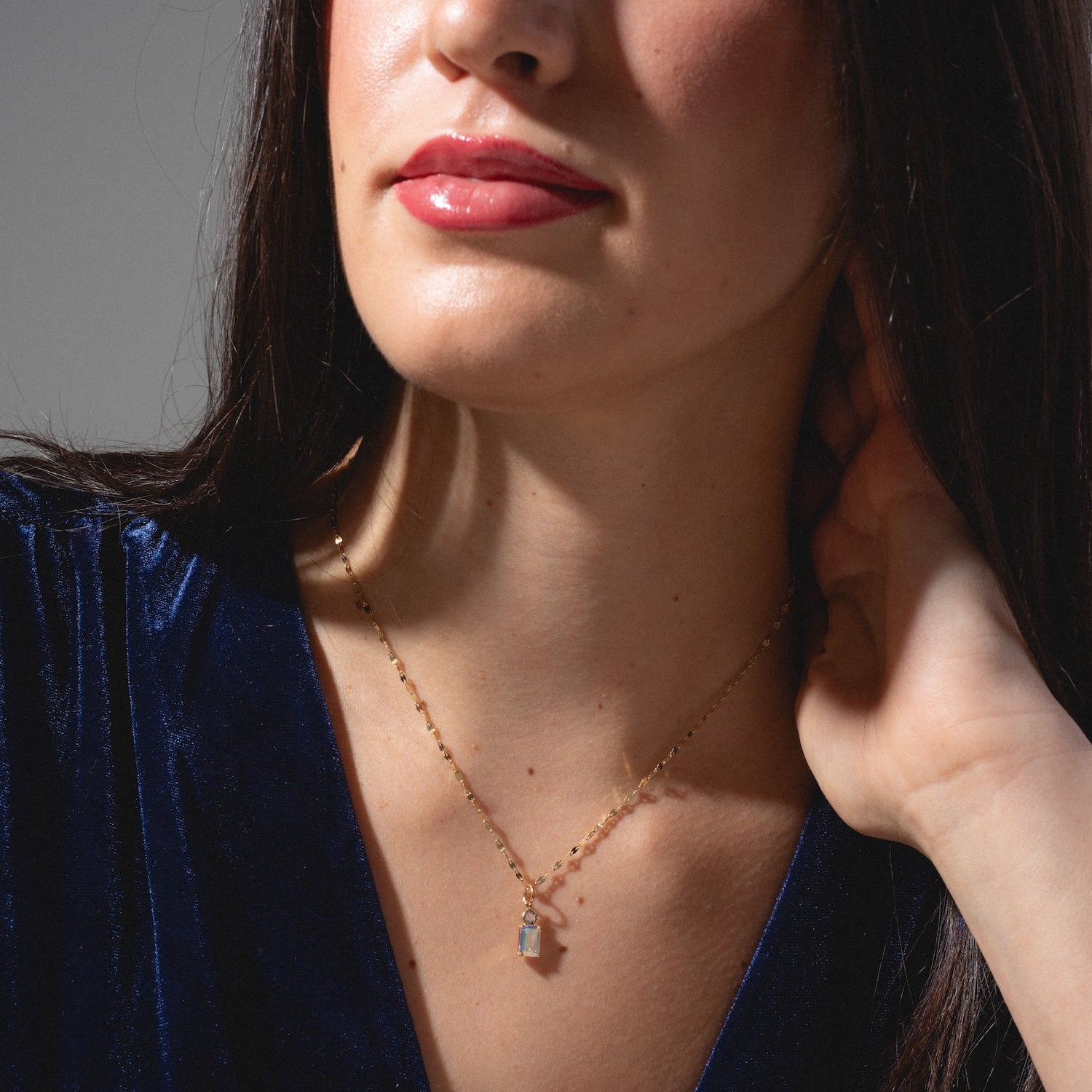 A woman with long dark hair wears a blue velvet top and the One of a Kind 14k Angel Aura and Diamond Charm necklace. She touches her hair with one hand, her glossy lips and partially visible face completing the look.