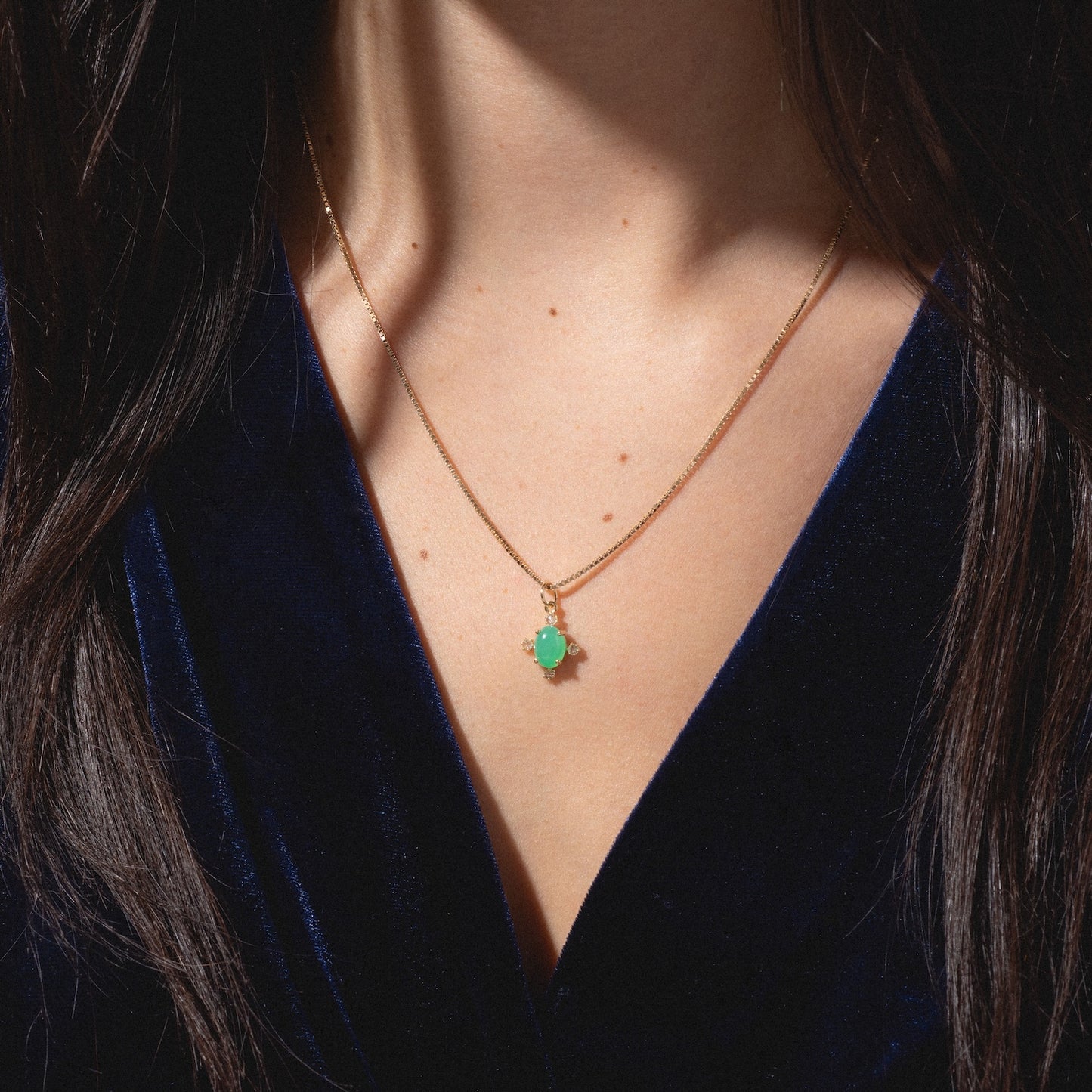 A woman's neck and chest adorned with the One of a Kind 14k Chrysoprase and Diamond Charm necklace, featuring a small green gemstone pendant, complement her deep blue velvet top as her dark hair frames the scene.
